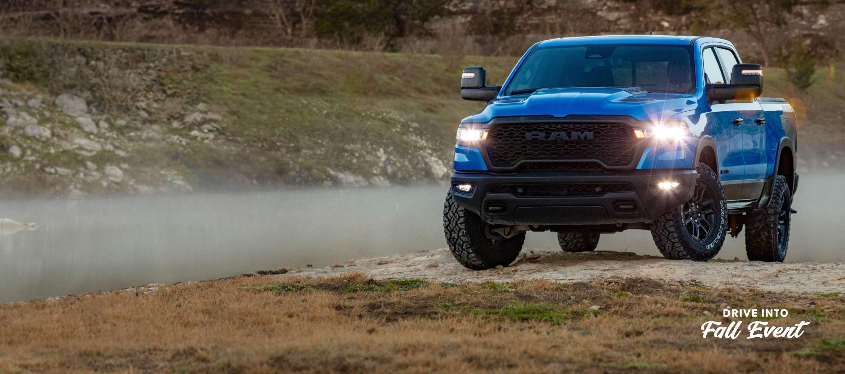 A blue 2025 Ram 1500 Rebel 4x4 Crew Cab with its headlamps and fog lamps on, traveling up a small hill off-road. Drive Into Fall Sales Event.