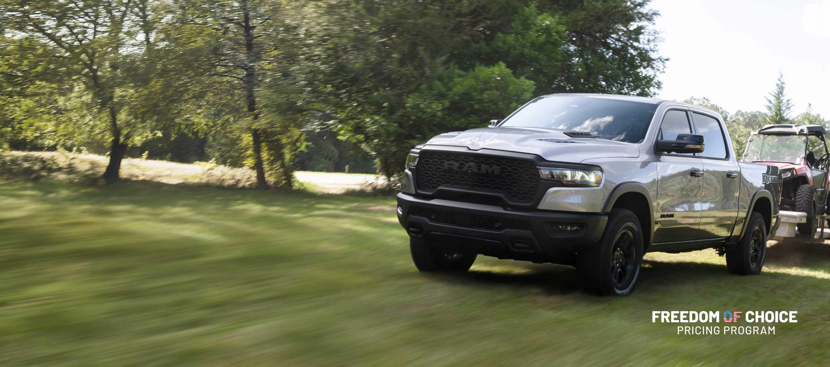 A silver 2025 Ram 1500 Rebel Crew Cab parked in a field, towing an ATV on a utility trailer. Truck Month. Freedom Of Choice Pricing Update Sales Event.