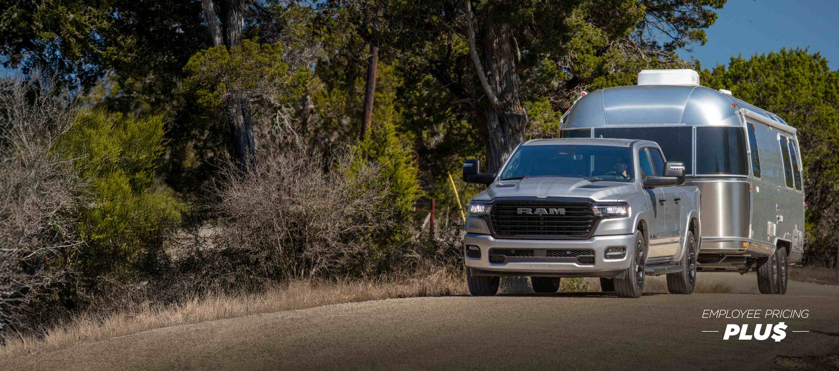 A silver 2025 Ram 1500 Tungsten Crew Cab being driven on a highway bridge in a city, with tall buildings in the background. Employee Pricing Plus Sales Event.