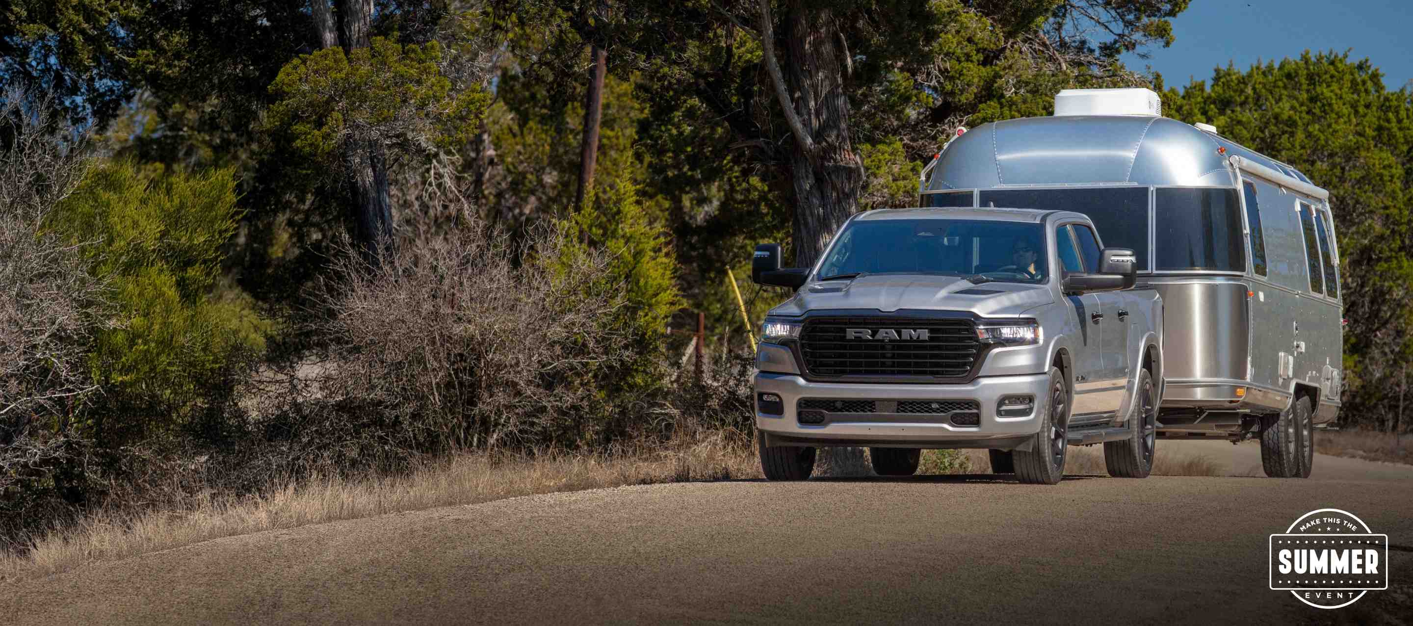 A silver 2025 Ram 1500 Tungsten Crew Cab being driven on a highway bridge in a city, with tall buildings in the background. Make This The Summer Event.