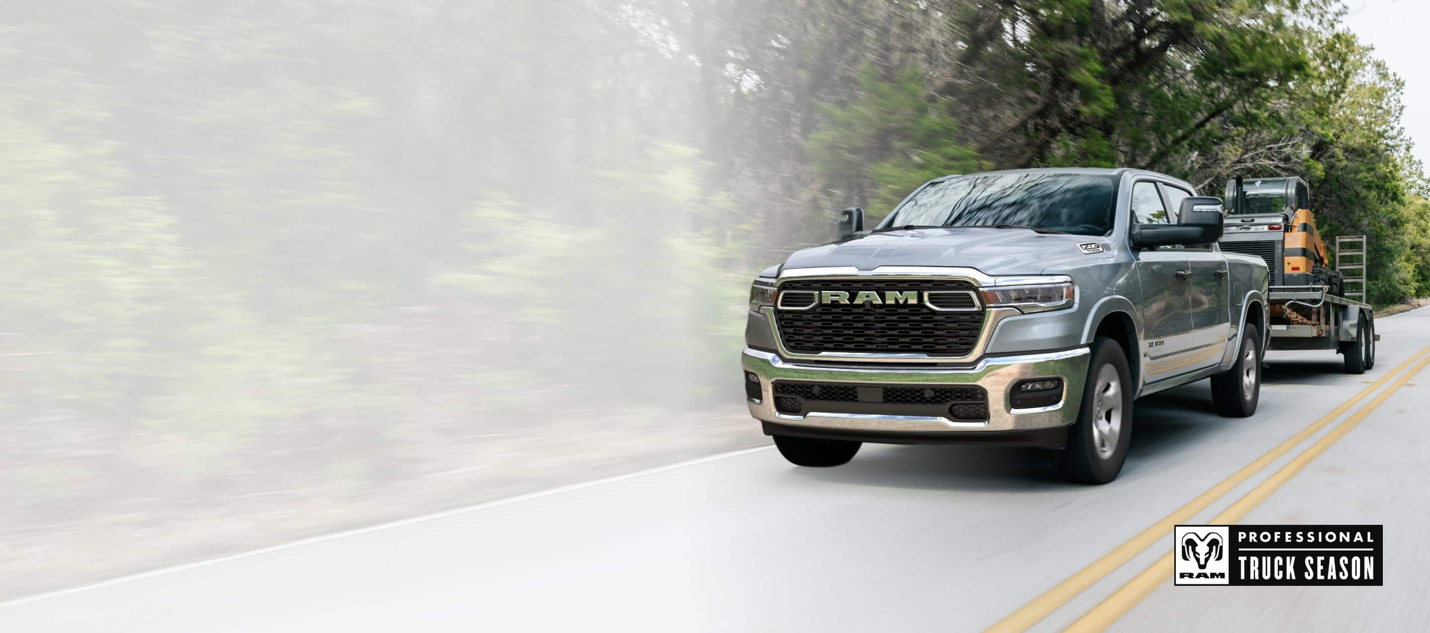A silver 2025 Ram 1500 Big Horn 4x4 Crew Cab towing motorized equipment as it travels down a highway. Ram Professional Truck Season.