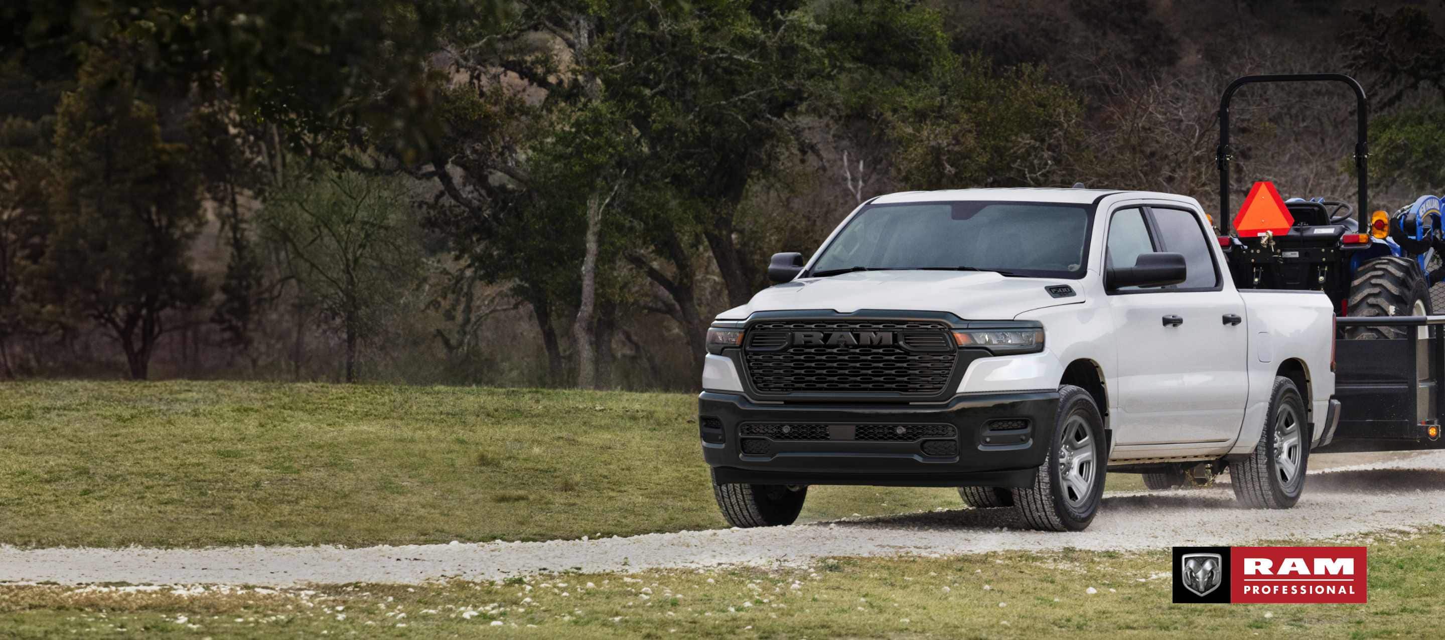 A white 2025 Ram 1500 Tradesman 4x4 Crew Cab towing a tractor on a landscape trailer. Ram Professional.