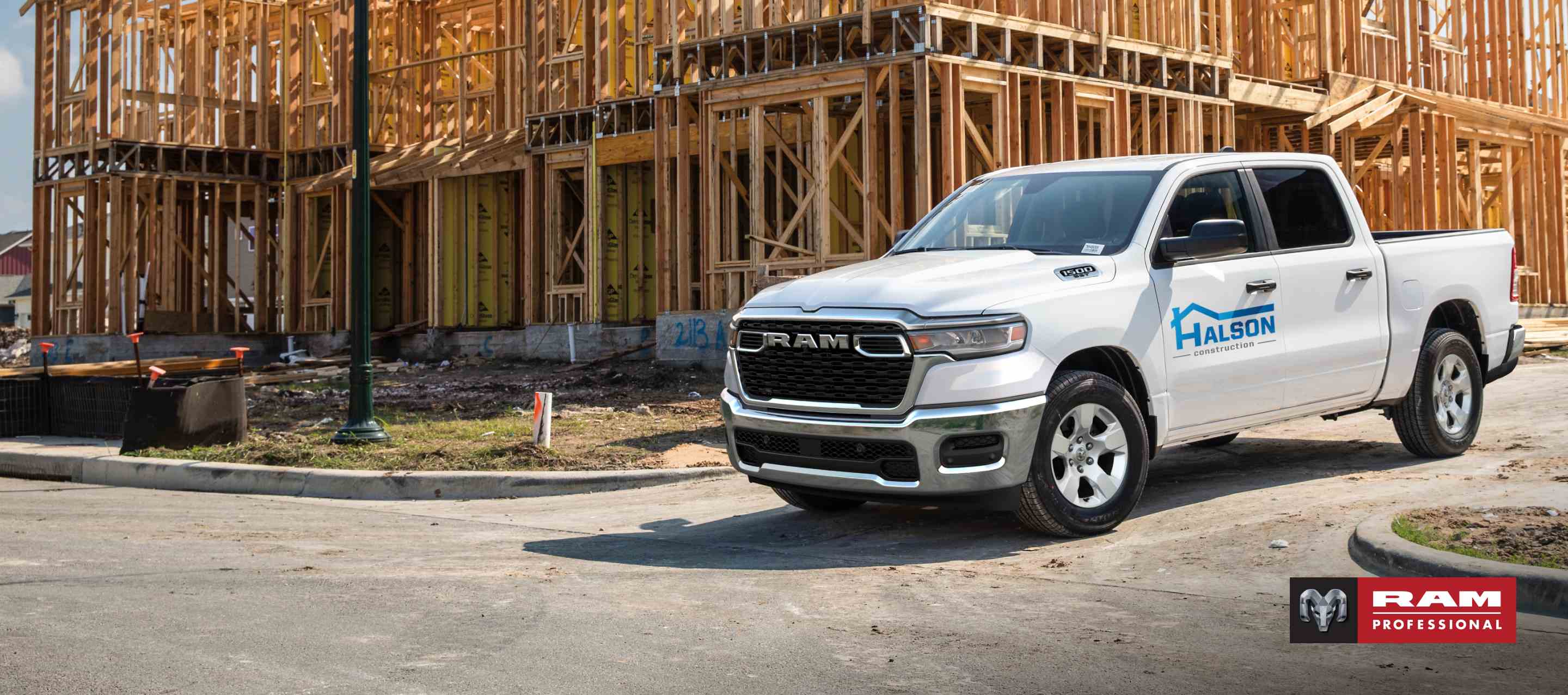 A white 2025 Ram 1500 Tradesman 4x4 Crew Cab with a construction company's signage on its driver's door, parked at a building site. Ram Professional.