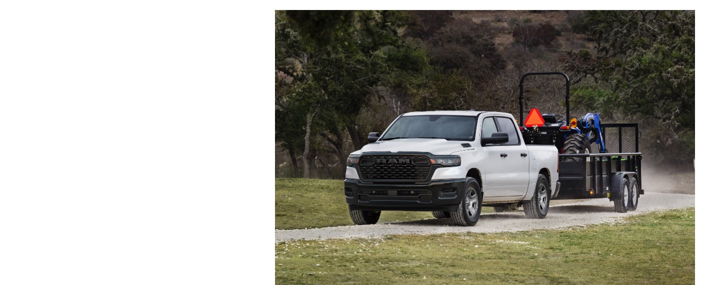 A white 2025 Ram 1500 Tradesman 4x4 Crew Cab towing a tractor on a landscape trailer.