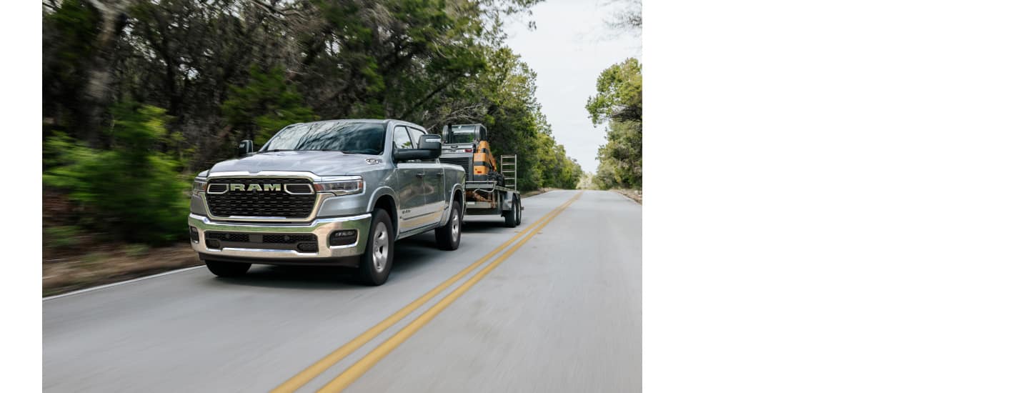 A silver 2025 Ram 1500 Big Horn 4x4 Crew Cab traveling on a highway, towing a construction vehicle on a tilt trailer.