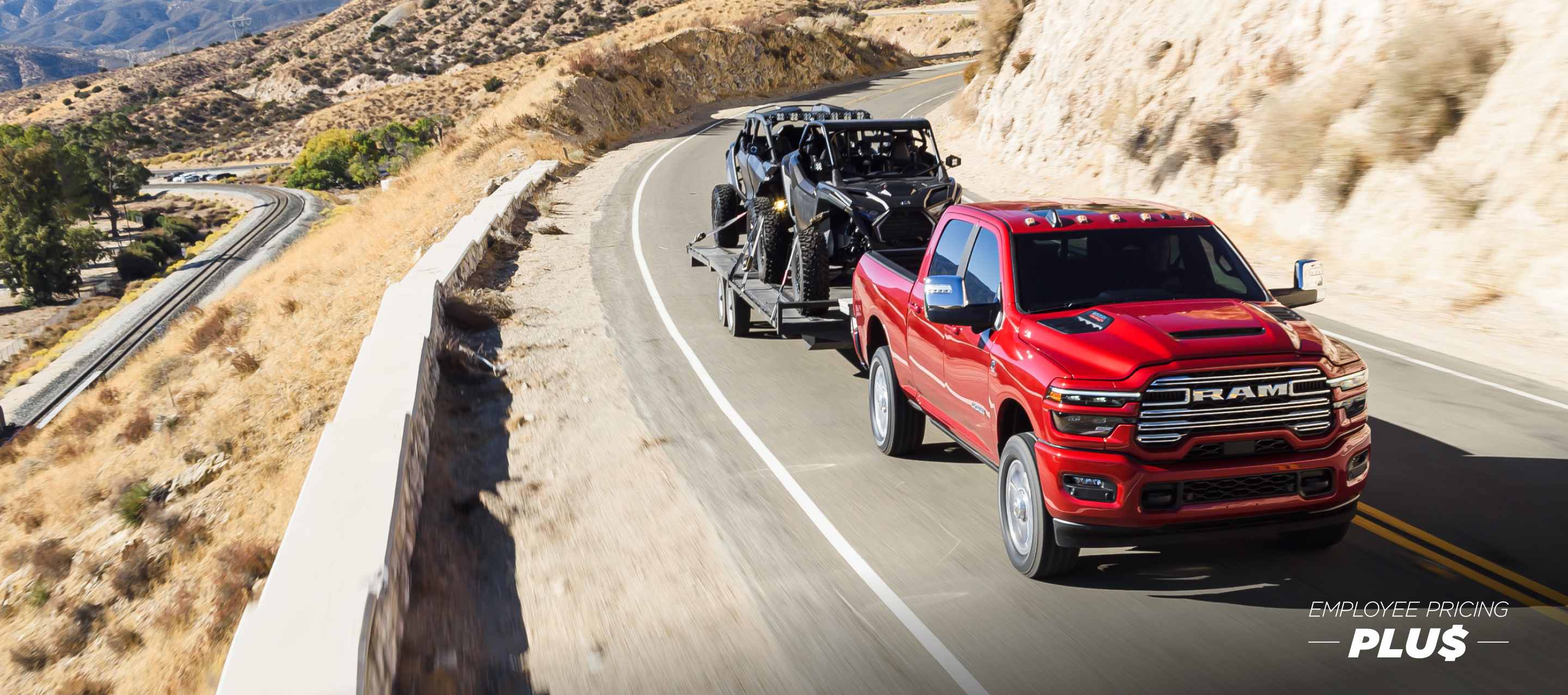 A red 2025 Ram 2500 Laramie 4x4 Crew Cab traveling on a highway in the mountains, while towing two ATVs. Employee Pricing Plus Sales Event.