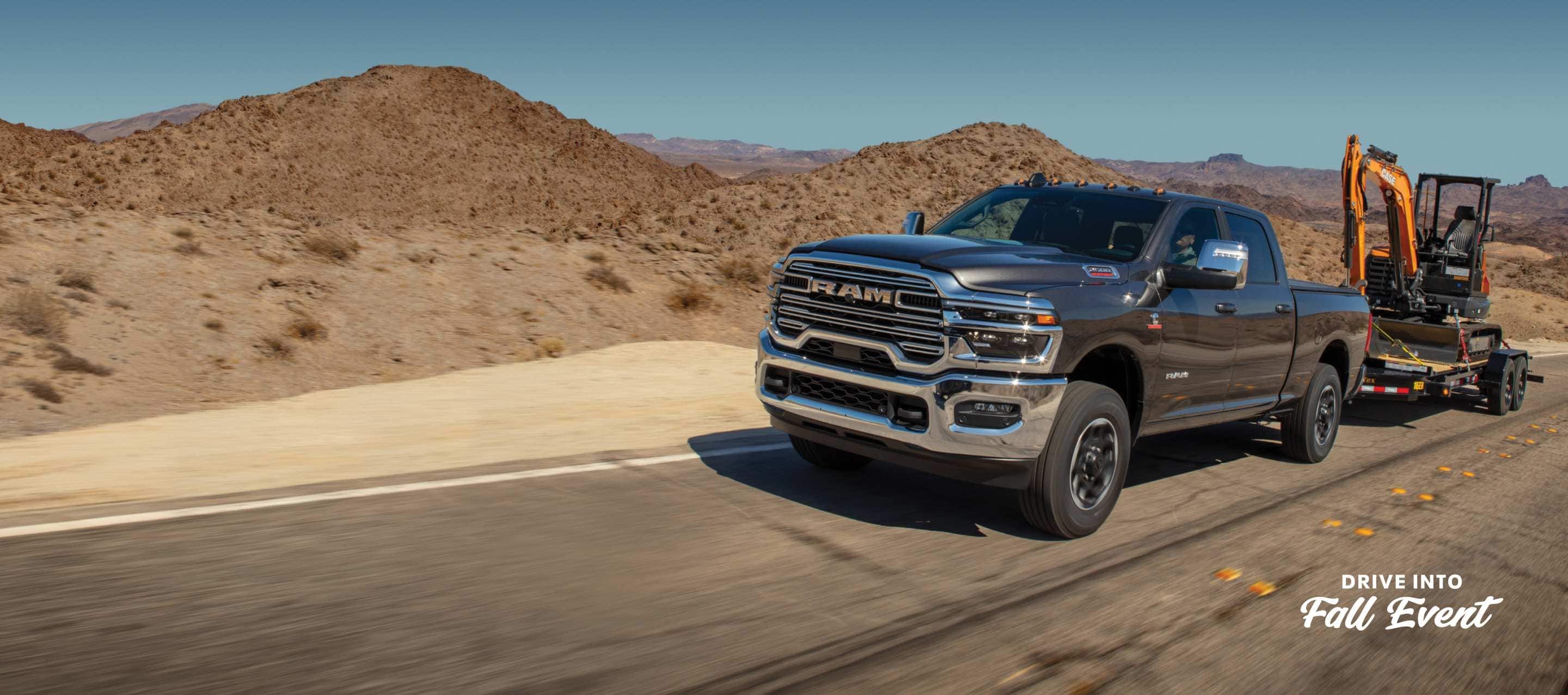 A gray 2025 Ram 2500 Laramie 4x4 Crew Cab traveling down a highway, towing an excavator on a flatbed trailer. Drive Into Fall Sales Event.
