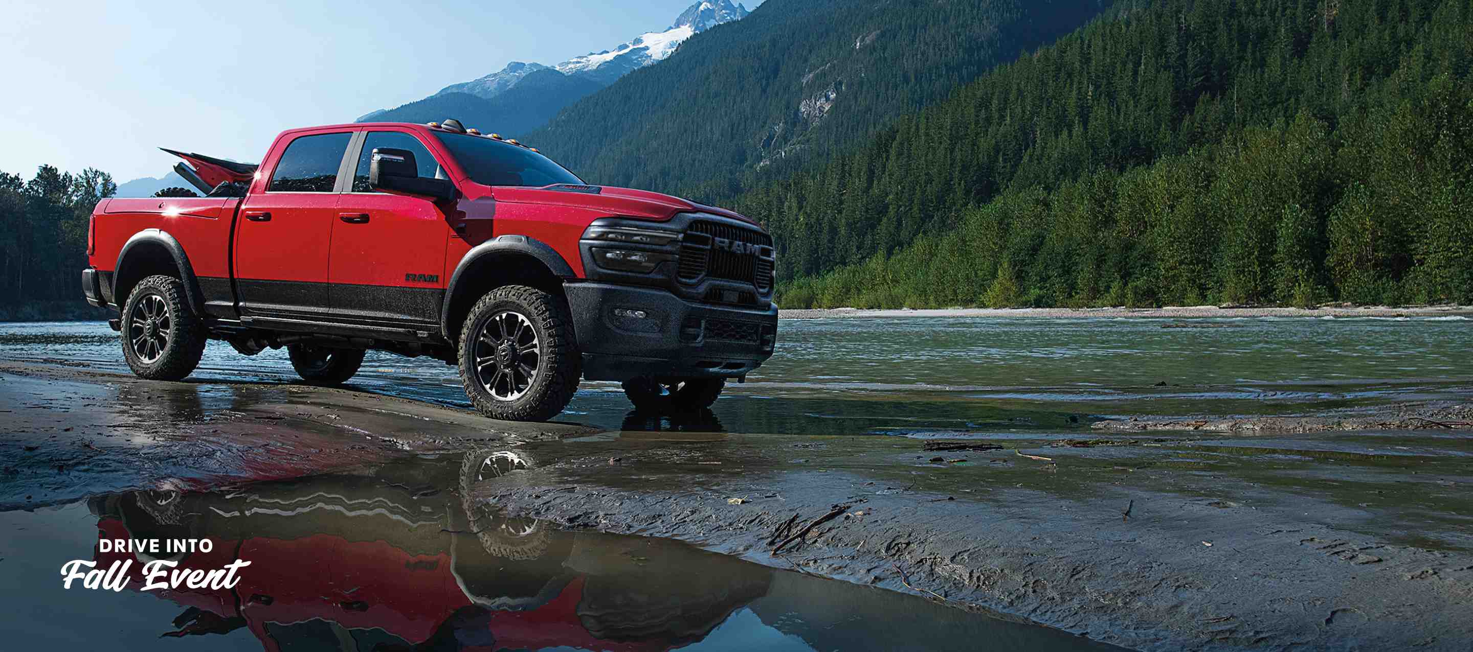 A red 2025 Ram 2500 Rebel 4x4 Crew Cab parked on a muddy trail beside a stream with mountains in the background. Drive Into Fall Sales Event.