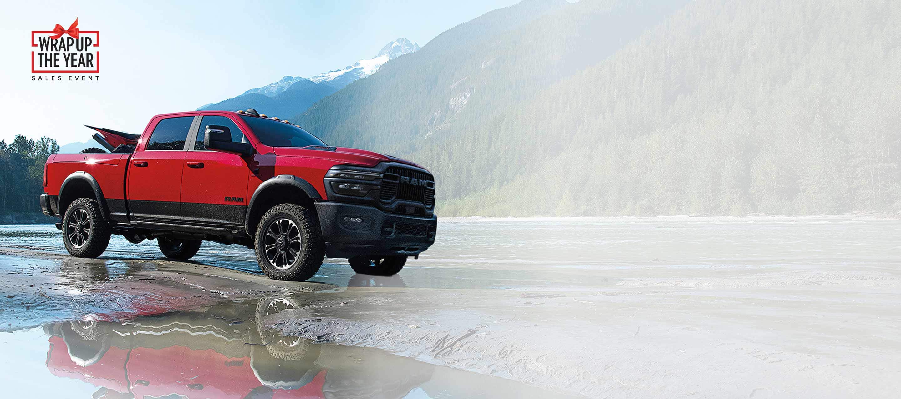 A red 2025 Ram 2500 Rebel 4x4 Crew Cab parked on a muddy trail beside a stream with mountains in the background. Wrap Up The Year Sales Event.