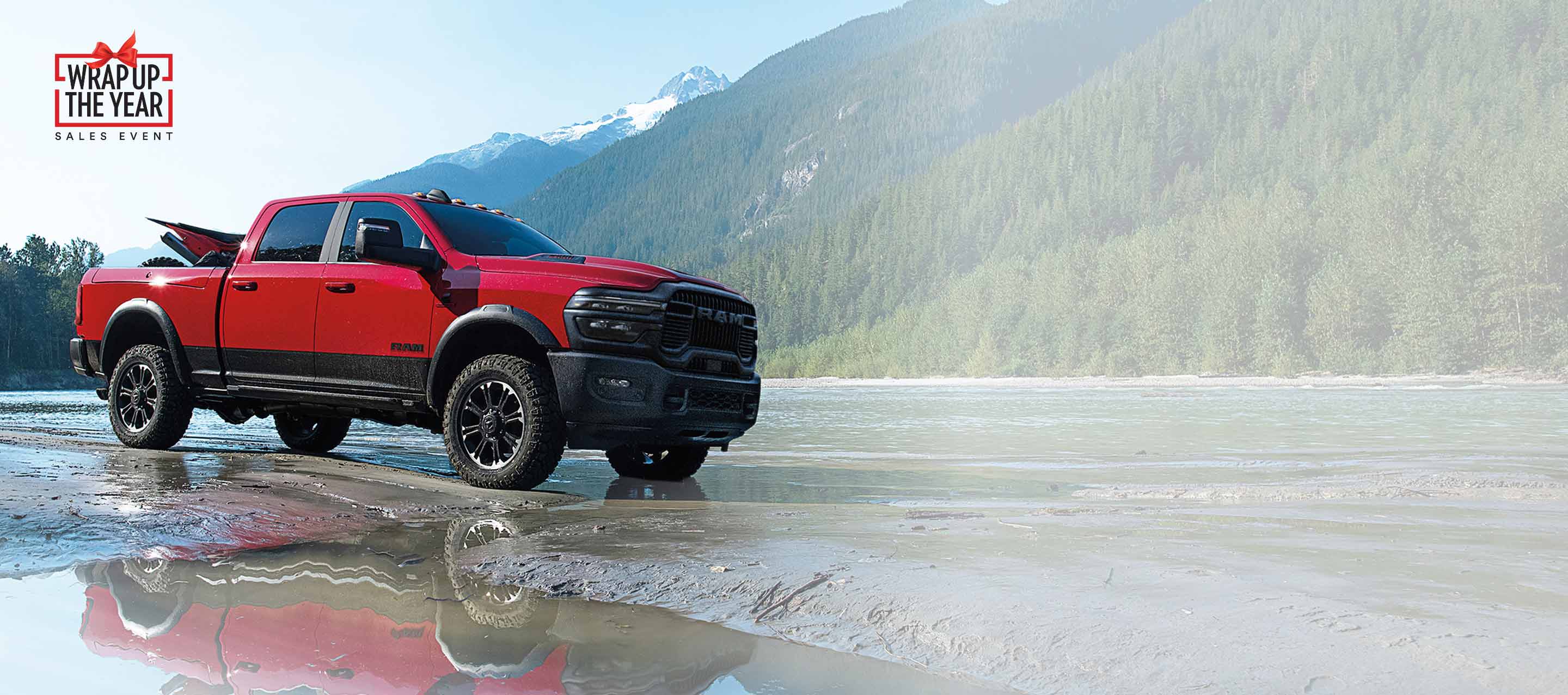 A red 2025 Ram 2500 Rebel 4x4 Crew Cab parked on a muddy trail beside a stream with mountains in the background. Wrap Up The Year Sales Event.