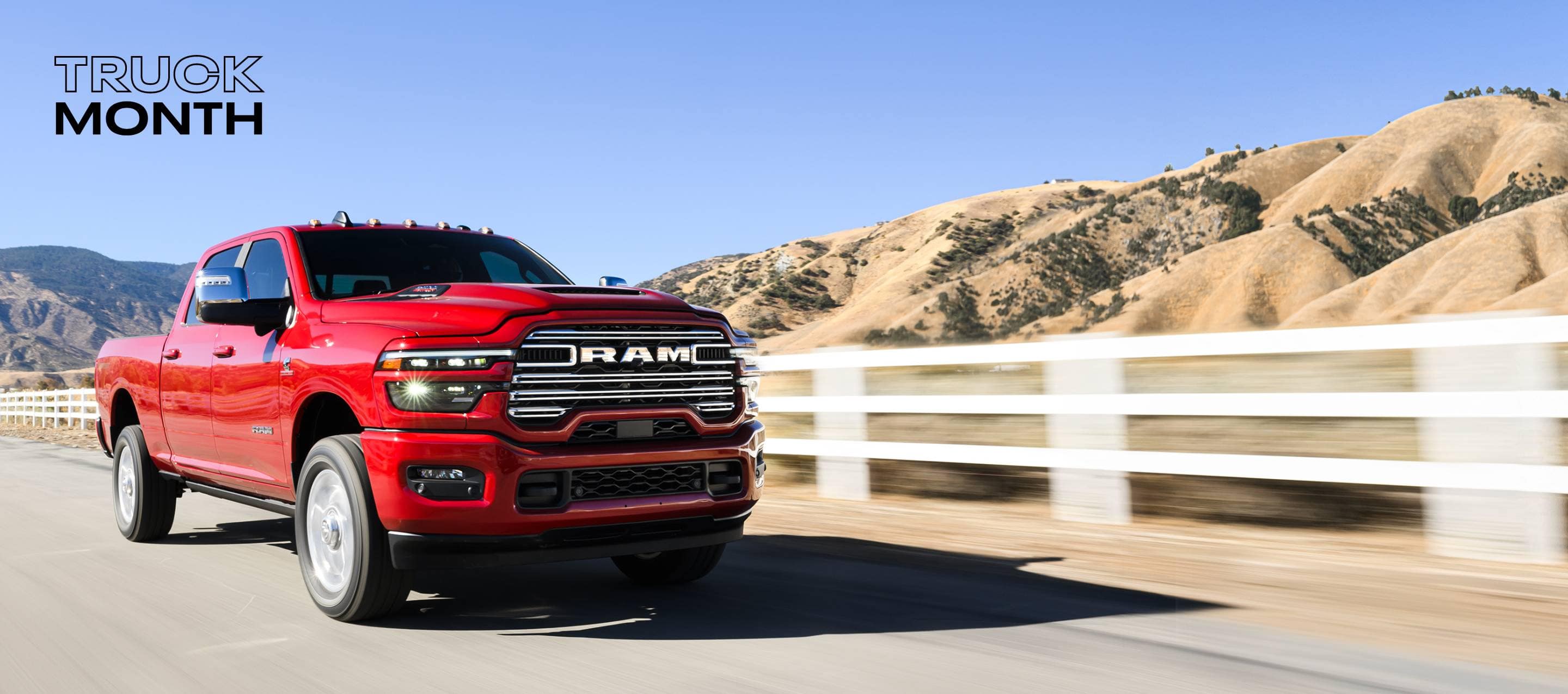 A red 2025 Ram 2500 Laramie 4x4 Crew Cab traveling on a highway with mountains in the background. Truck Month.