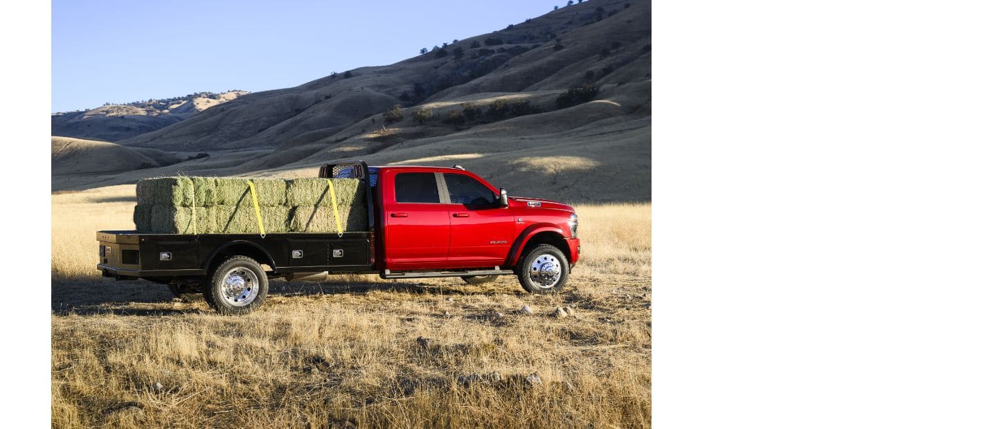 A passenger-side profile of a red 2025 Ram 5500 Laramie Chassis Crew Cab in a field, carrying several large bales of hay on a long platform upfit.