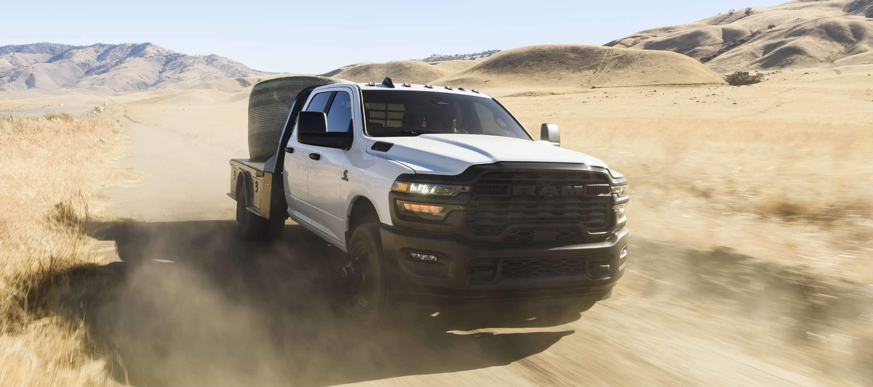 A white 2025 Ram 3500 Tradesman Chassis Crew Cab traveling through a desert with a huge bale of hay on its platform upfit.
