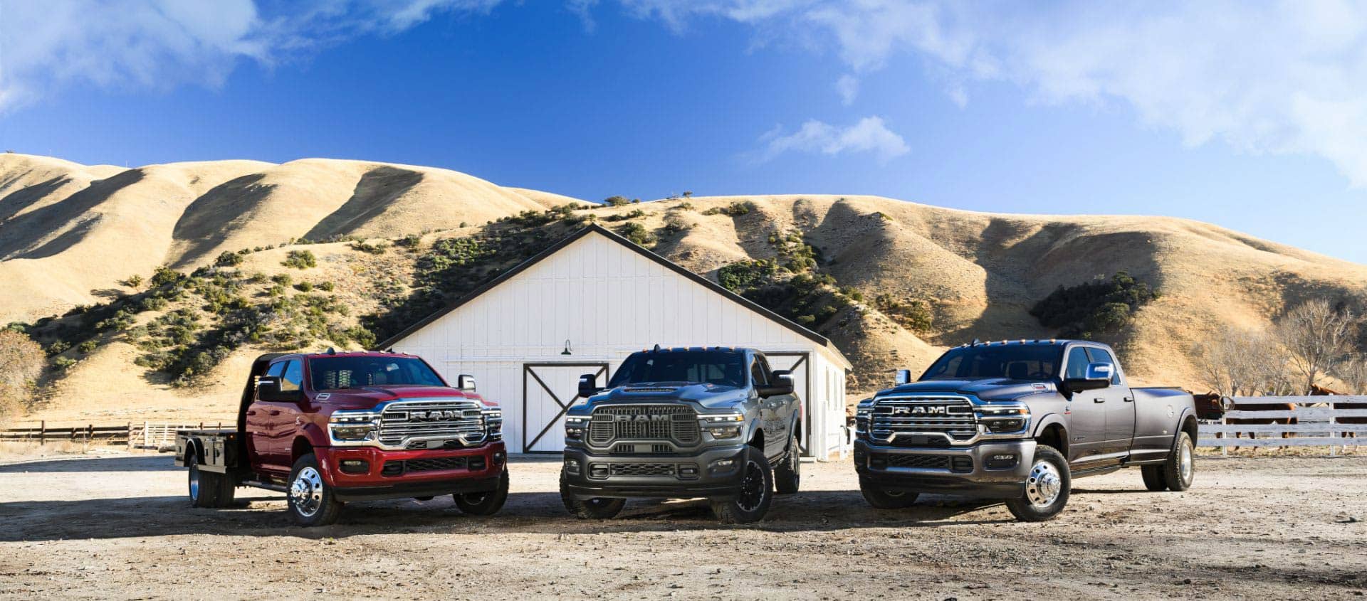 Three Ram trucks parked at a ranch. From left to right: a red 2025 Ram 5500 Laramie Chassis Crew Cab, a blue 2025 Ram 2500 Rebel Crew Cab and a gray 2025 Ram 3500 Limited Longhorn Crew Cab.