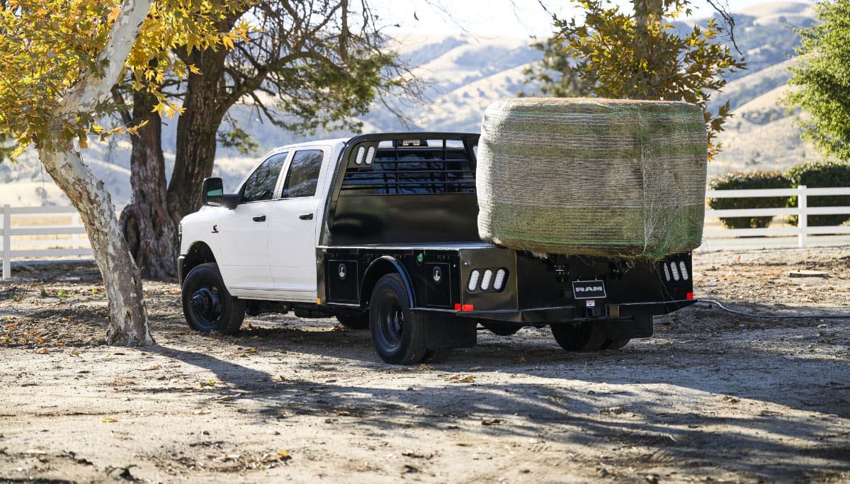 Display A white 2025 Ram 3500 Tradesman Chassis Crew Cab with a huge bale of hay on its platform upfit, parked at a farm.