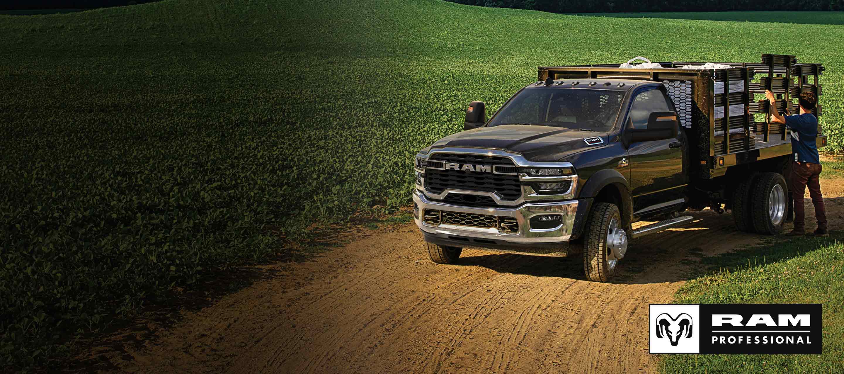 A black 2025 Ram 5500 Tradesman Chassis Cab Regular Cab with a landscaping upfit, parked in a large farmer's field with a man adjusting one of the upfit side panels. Ram Professional.