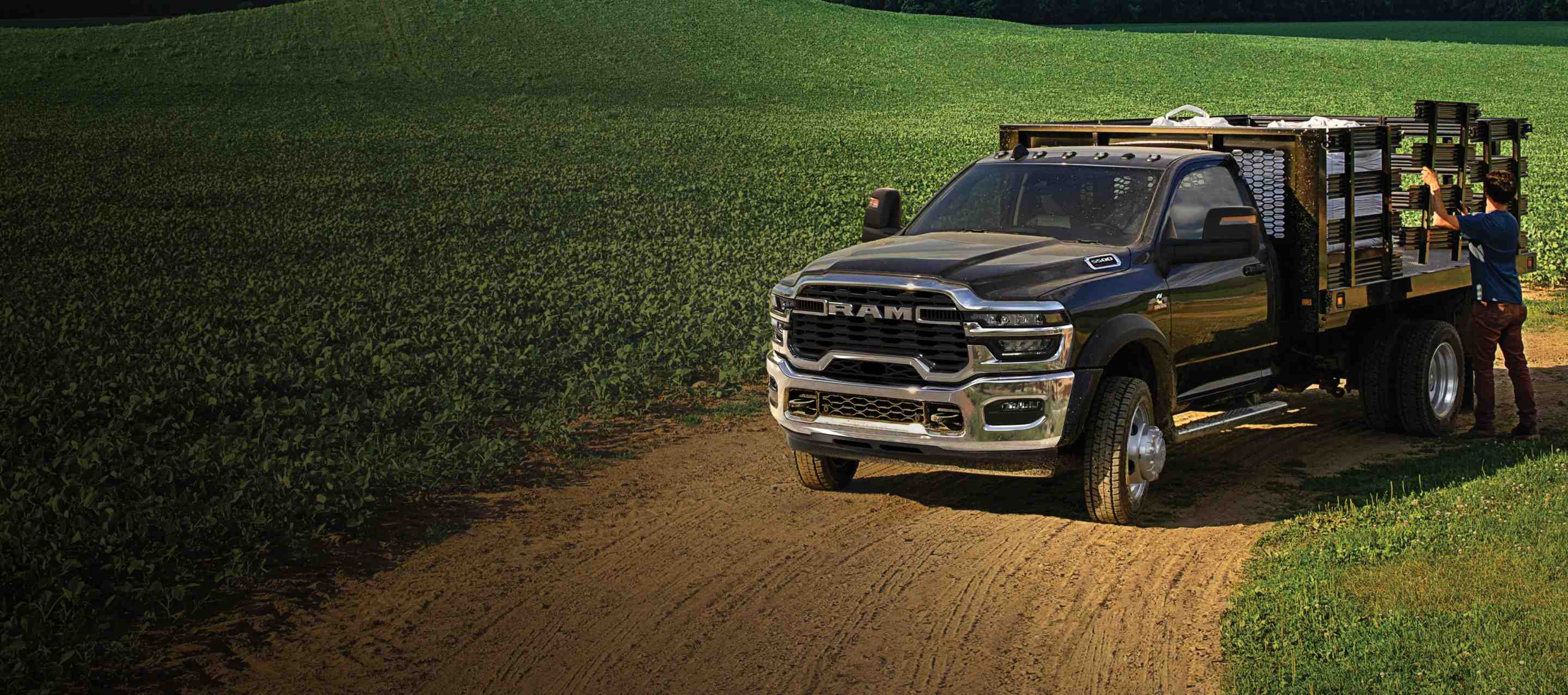 A black 2025 Ram 5500 Tradesman Chassis Cab Regular Cab with a landscaping upfit, parked in a large farmer's field with a man adjusting one of the upfit side panels. Ram Professional.
