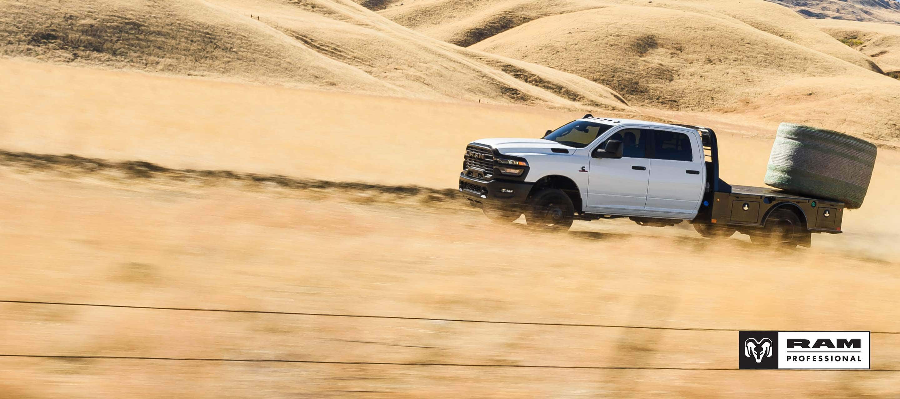 A white 2025 Ram 3500 Tradesman Crew Cab Chassis Cab climbing a sand dune in the desert, with a huge bale of hay on its platform upfit. Ram Professional SALES EVENT: Ram Professional