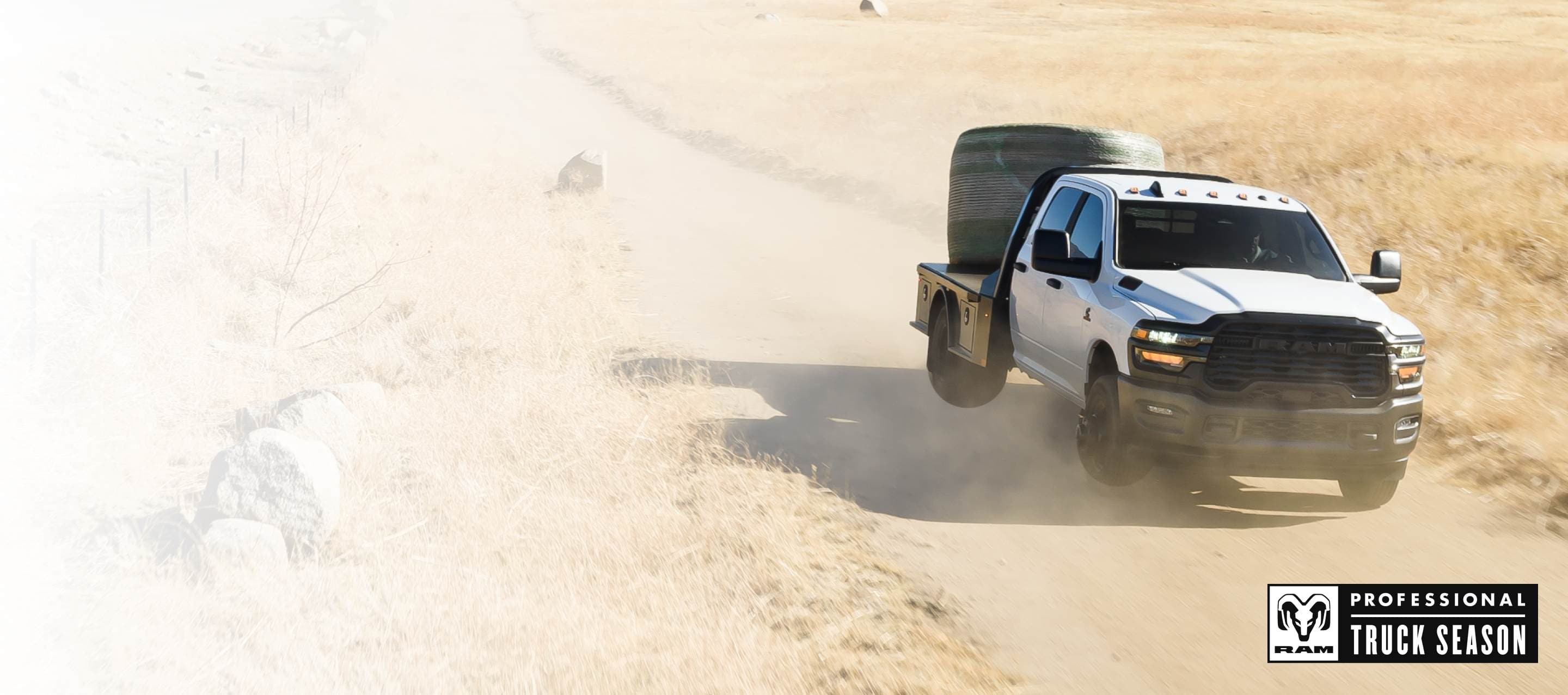 A white 2025 Ram 3500 Tradesman 4x4 Chassis Cab Crew Cab traveling through cattle country, carrying a bale of hay on a platform upfit. Ram Professional Truck Season.