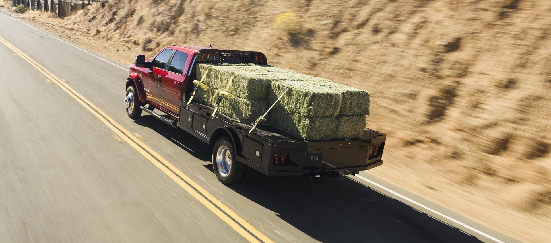 A red 2025 Ram 5500 Laramie Chassis Crew Cab traveling down a highway, carrying several large bales of hay on a long platform upfit.