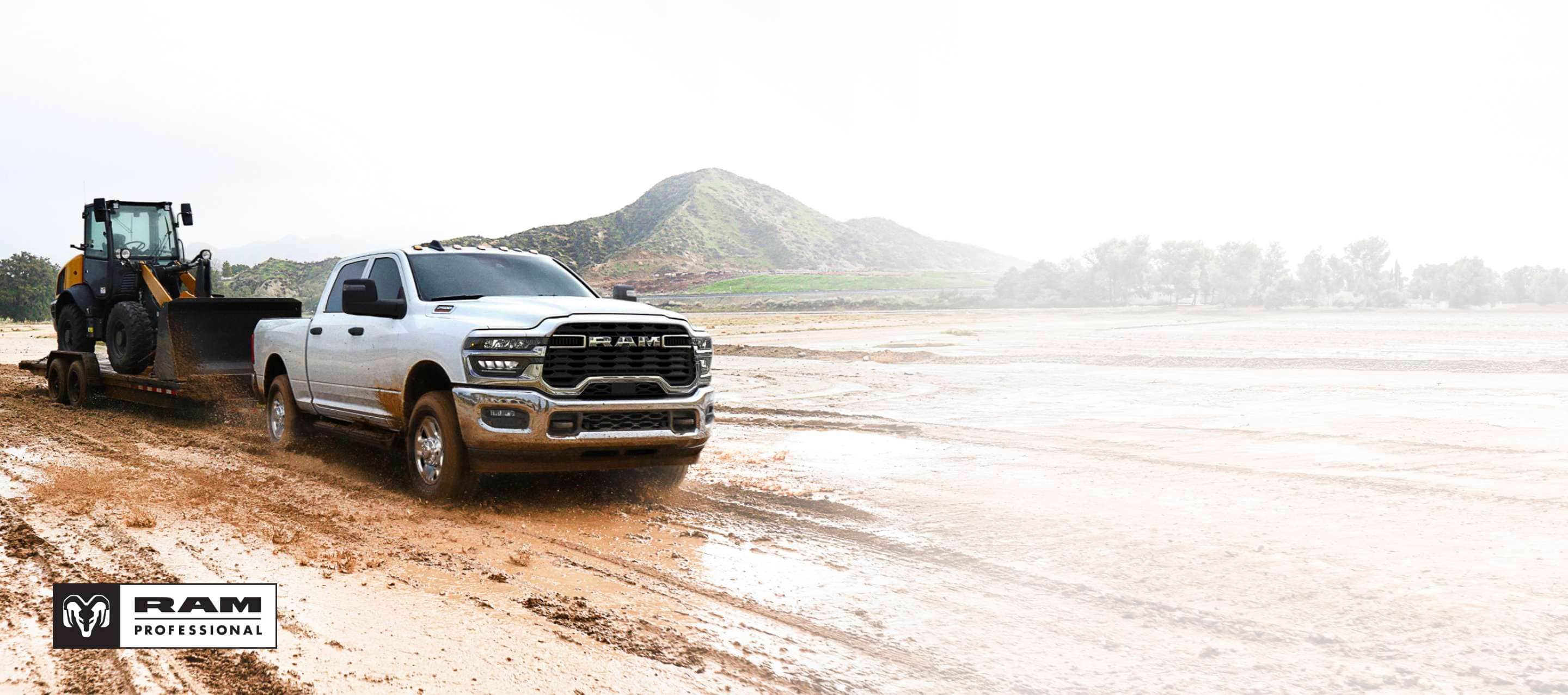 A white 2025 Ram 2500 Tradesman 4x4 Crew Cab towing a front end loader on a flatbed trailer through a muddy construction site. Ram Professional.