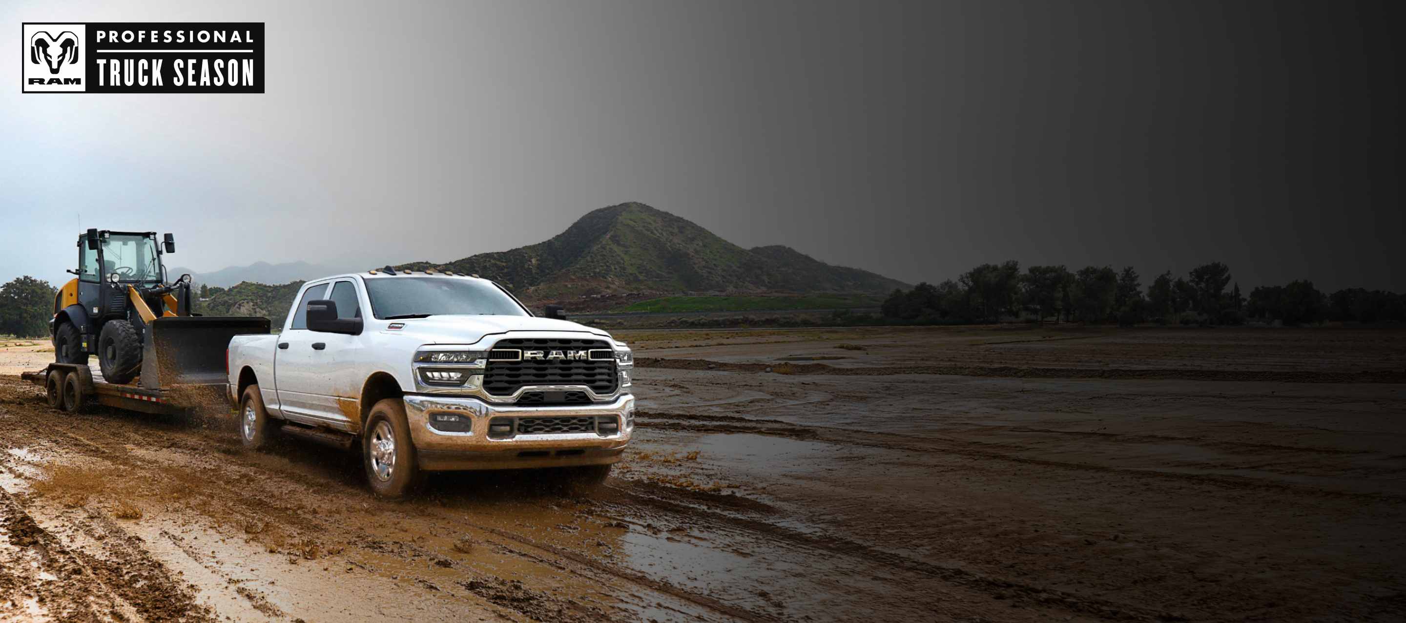 A white 2025 Ram 2500 Tradesman 4x4 Crew Cab towing a front end loader on a flatbed trailer through a muddy construction site. Ram Professional Truck Season.