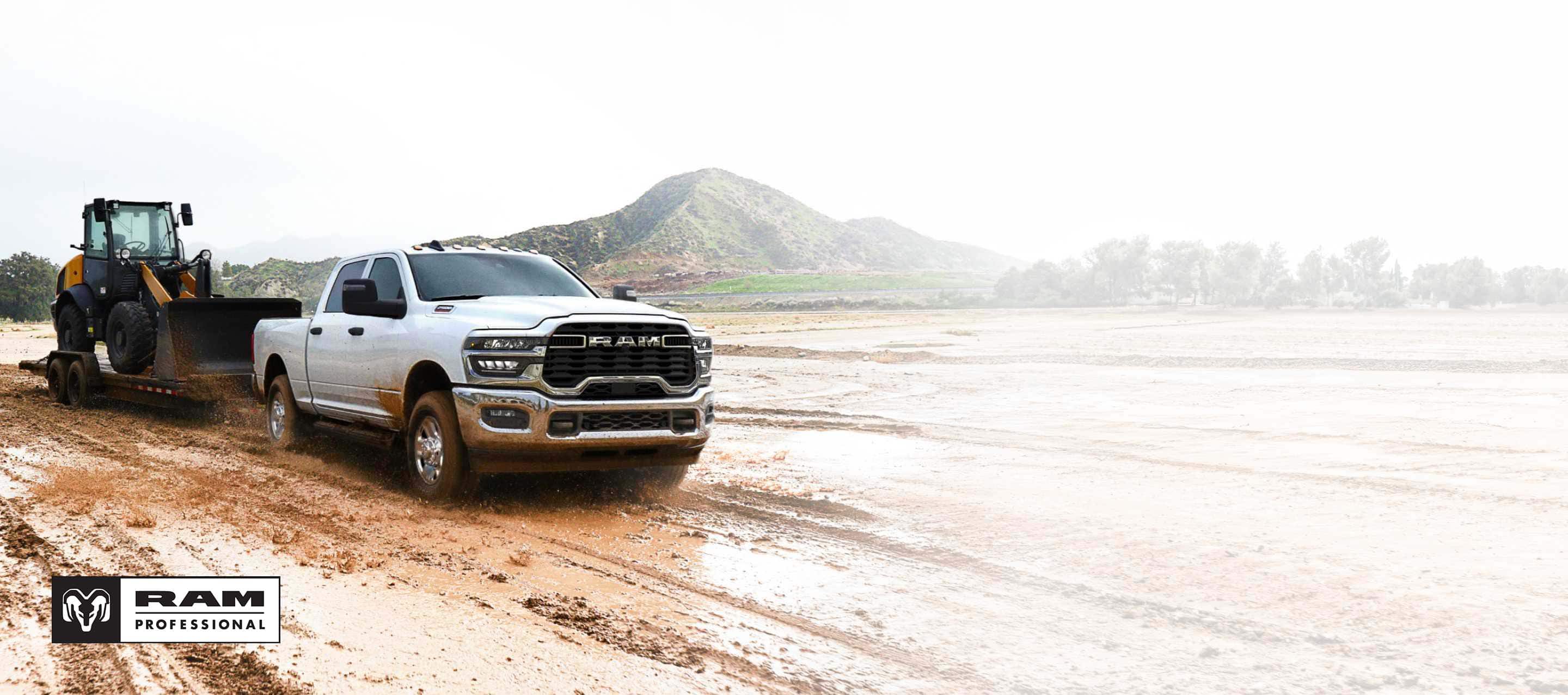 A white 2025 Ram 2500 Tradesman 4x4 Crew Cab towing a front end loader on a flatbed trailer through a muddy construction site. Ram Professional.