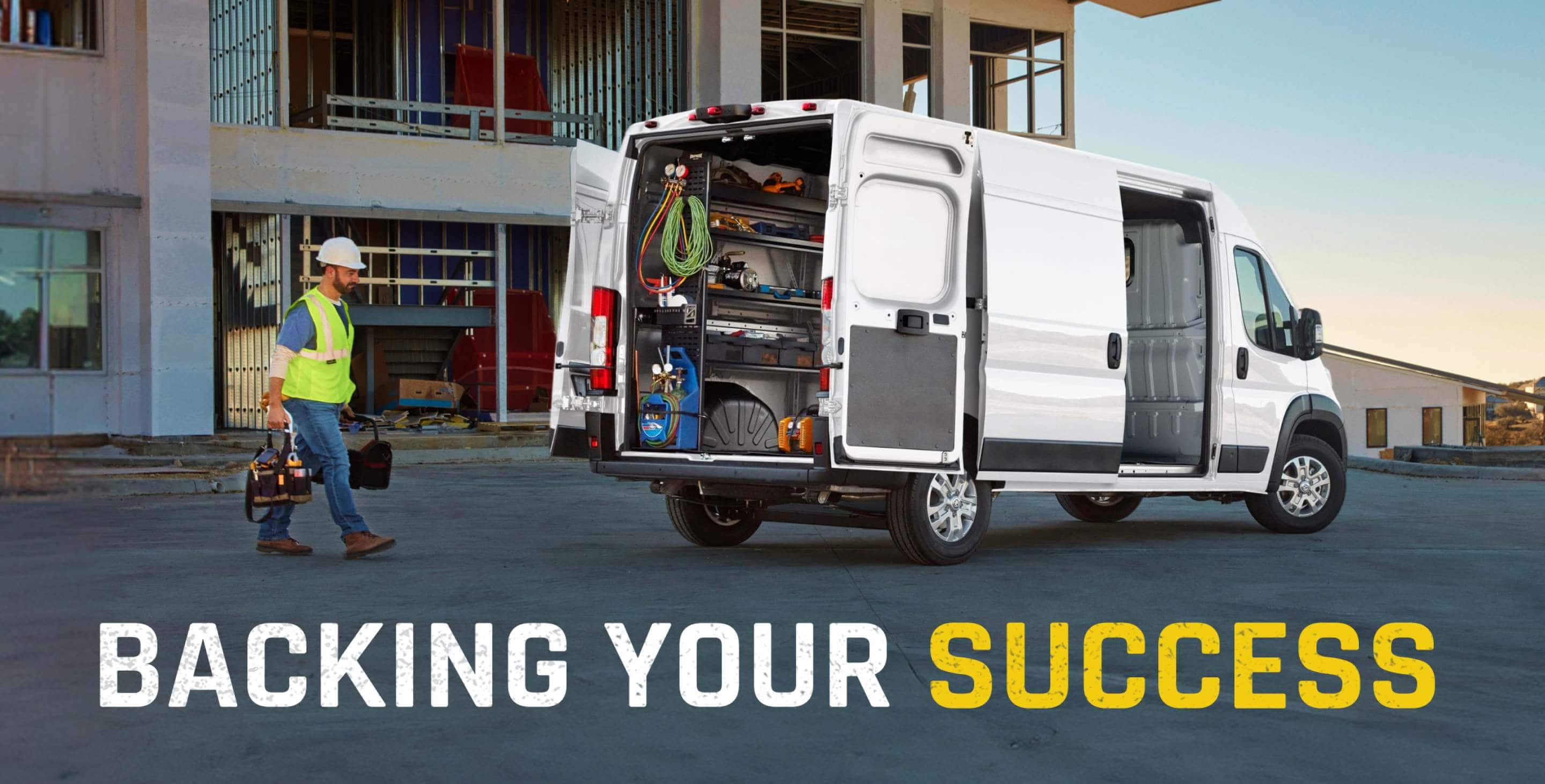 The rear of a white 2026 Ram ProMaster 3500 SLT Cargo Van High Roof with its passenger-side sliding door and rear doors open, displaying shelves full of tools and a technician walking toward the van.