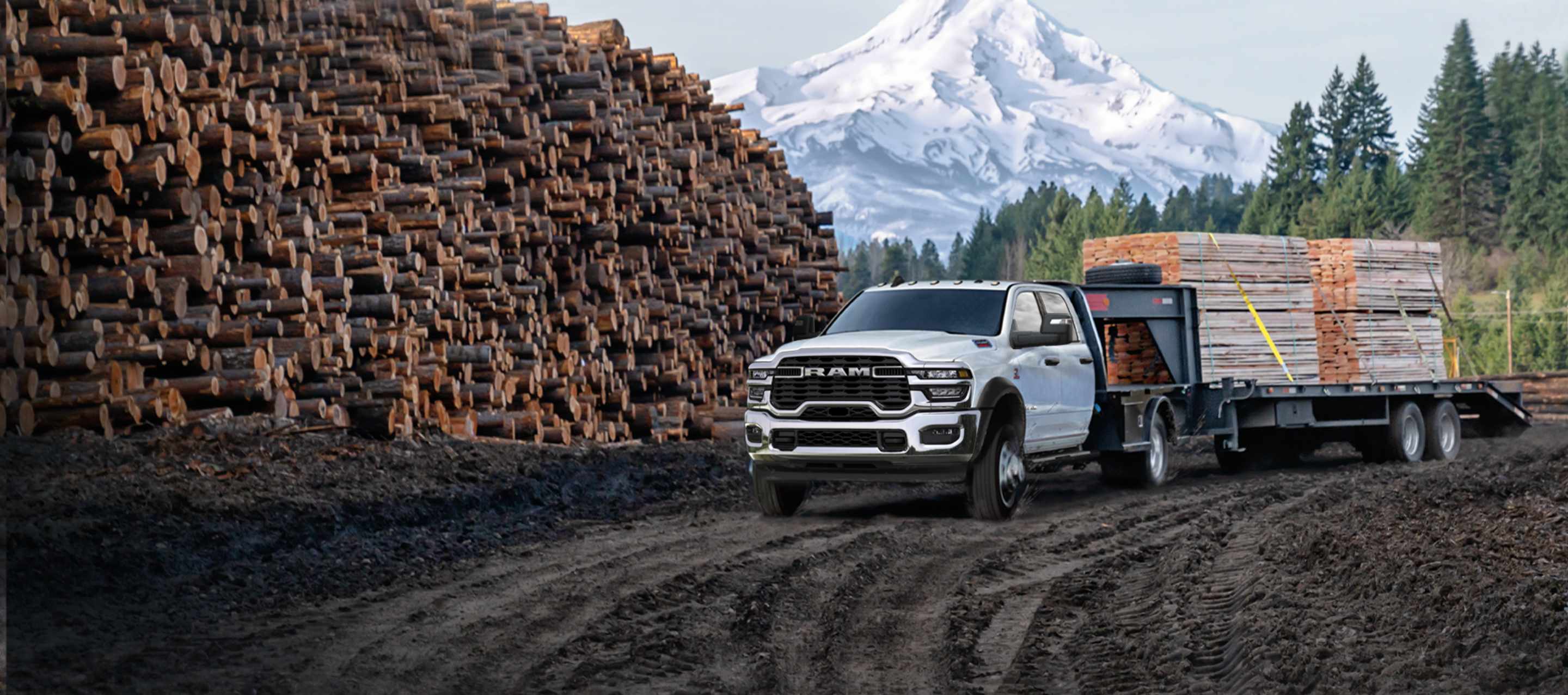 A 2025 Ram Tradesman Chassis Cab Crew Cab with a gooseneck trailer filled with lumber, parked beside a forest and huge mound of cut logs, with a snow-capped mountain in the background.