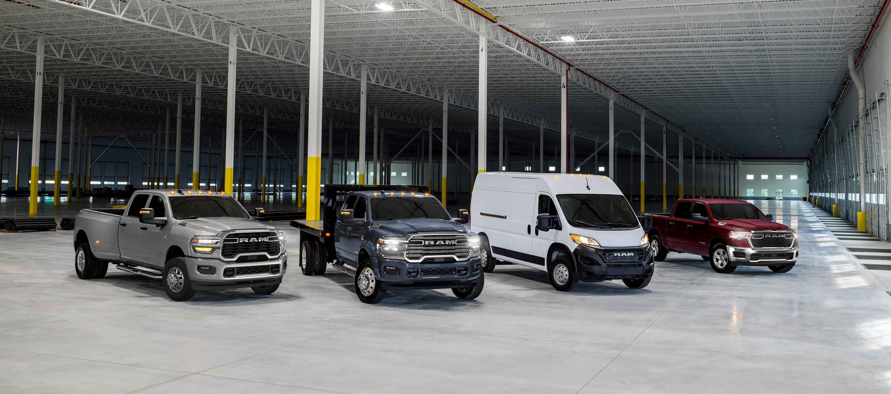 A lineup of four 2025 Ram Brand vehicles in an industrial garage. From left to right: a silver Ram 3500 Big Horn 4x4 Crew Cab, a blue Ram 5500 Laramie 4x4 Chassis Cab Crew Cab with utility bed upfit, a white Ram ProMaster 2500 Cargo Van High Roof and a red Ram 1500 Tradesman 4x4 Crew Cab.