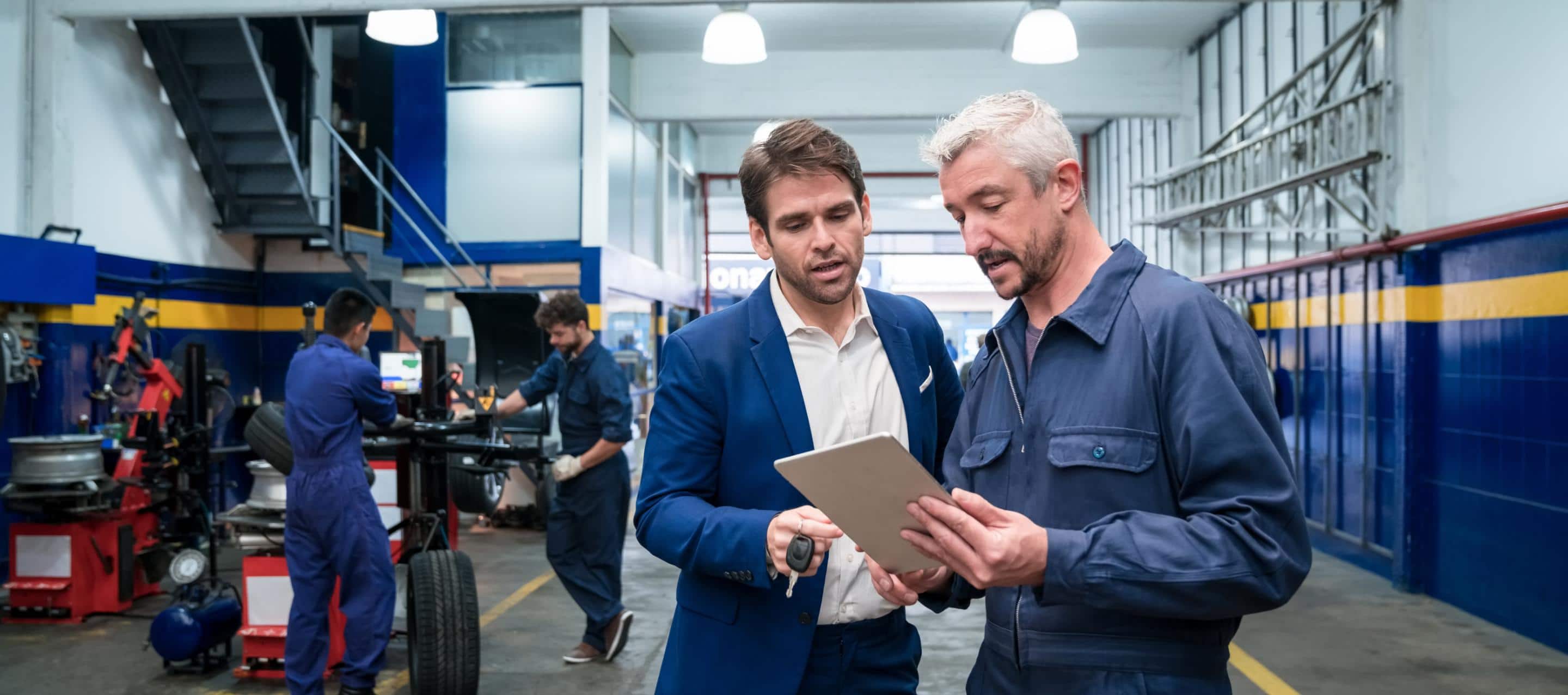 The service department in a dealership with a service advisor talking to a customer in the foreground and technicians working in the background.
