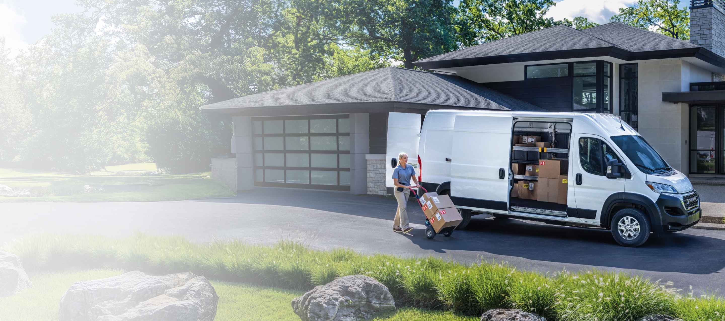 A white 2026 Ram ProMaster EV 3500 SLT High Roof Cargo Van with its passenger-side sliding door and rear doors open, revealing packages inside, parked in the driveway of a neighborhood home, with a delivery person nearby pushing a dolly full of boxes.