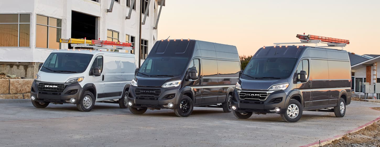 A lineup of three 2025 Ram ProMaster vans, each with a different roof height, parked at a construction site: from left to right, a white Ram ProMaster 1500 Tradesman Cargo Van Standard Roof, a black Ram ProMaster 3500 SLT Plus Cargo Van Super High Roof and a black Ram ProMaster 2500 SLT Cargo Van High Roof.