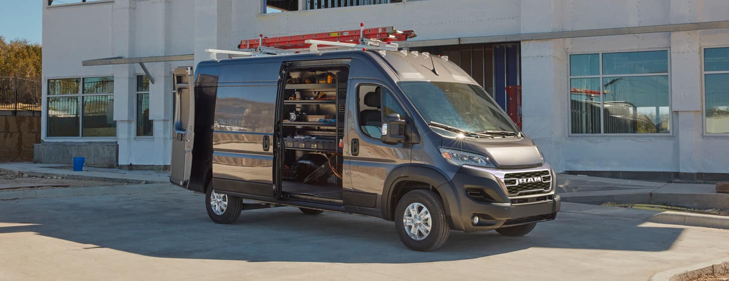 A passenger-side profile of a gray 2025 Ram ProMaster 3500 SLT Plus Cargo Van High Roof parked beside a commercial building with a ladder on its roof rack, its rear doors and sliding side door open revealing shelving full of tools in the cargo area.