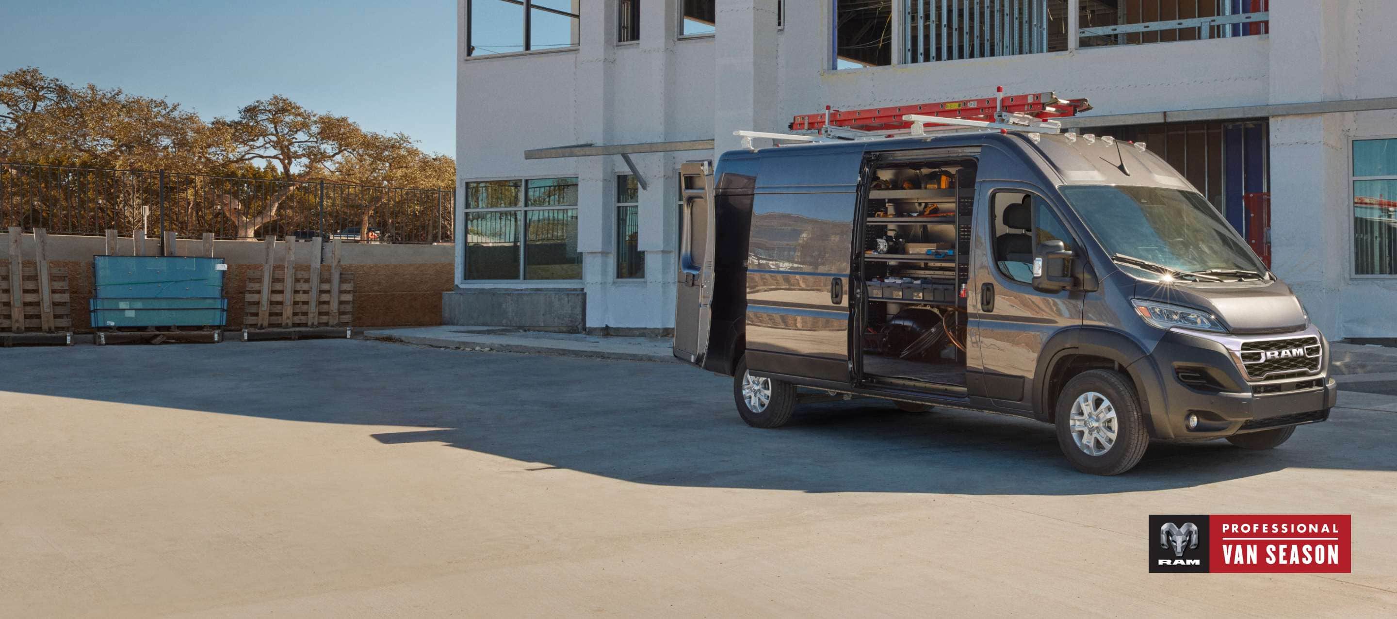A passenger-side profile of a gray 2025 Ram ProMaster 3500 SLT Plus High Roof Cargo Van parked beside a commercial building with a ladder on its roof rack, its rear doors and sliding side door open revealing shelving full of tools in the cargo area. Professional Van Season.