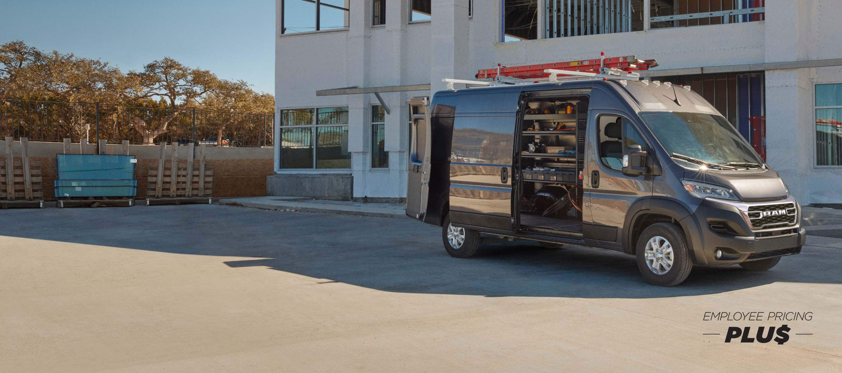 A passenger-side profile of a gray 2025 Ram ProMaster 3500 SLT Plus High Roof Cargo Van parked beside a commercial building with a ladder on its roof rack, its rear doors and sliding side door open revealing shelving full of tools in the cargo area. Employee Pricing Plus Sales Event.