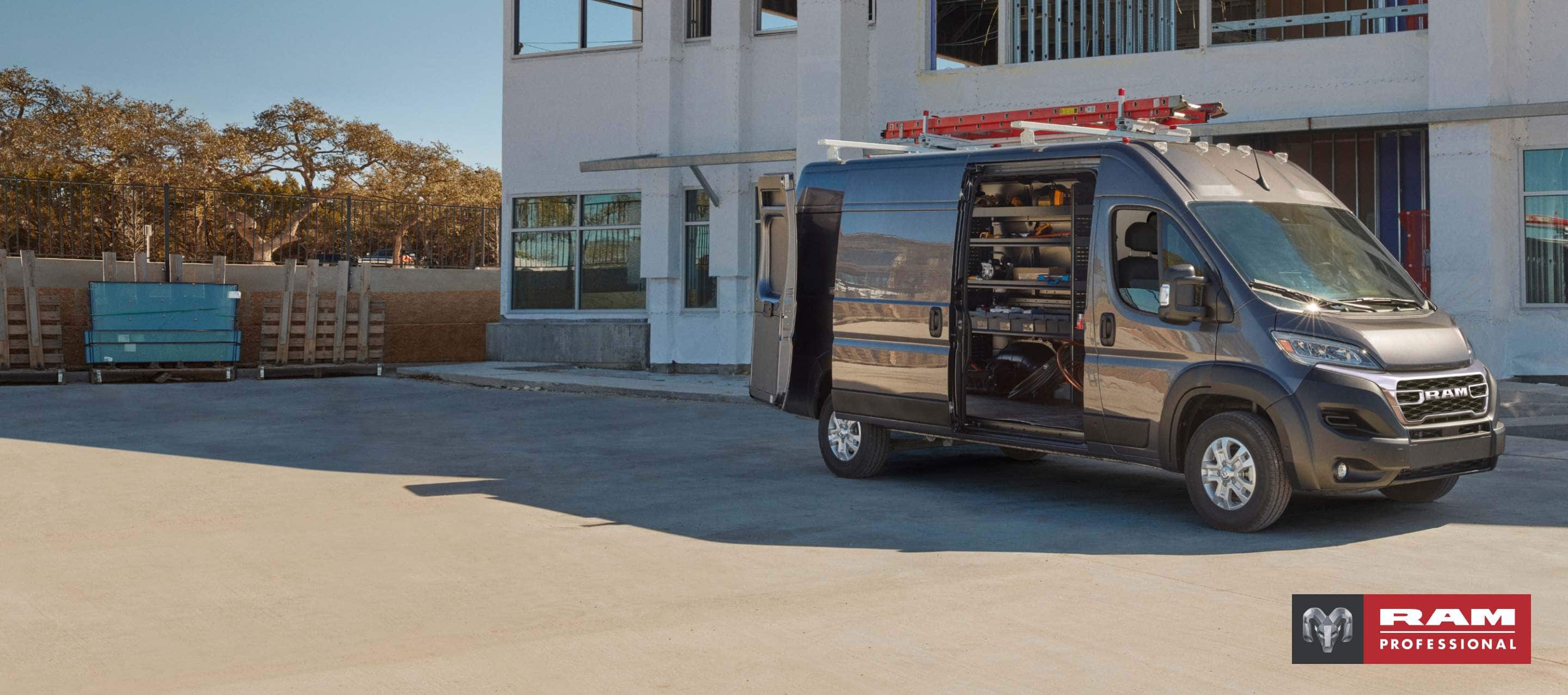 A passenger-side profile of a gray 2025 Ram ProMaster 3500 SLT Plus High Roof Cargo Van parked beside a commercial building with a ladder on its roof rack, its rear doors and sliding side door open revealing shelving full of tools in the cargo area. Ram Professional.