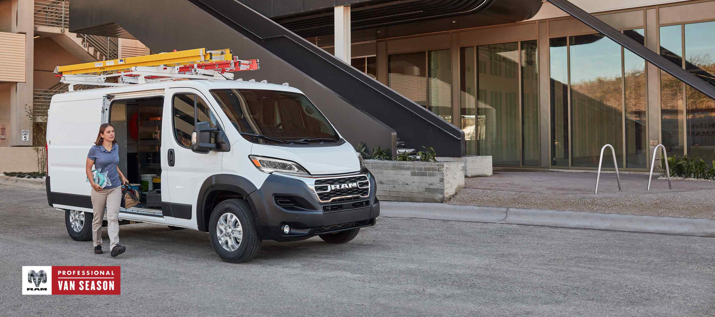 A passenger-side front angle of a white 2025 Ram ProMaster SLT Standard Roof Cargo Van, parked next to a contemporary building with two ladders on its roof rack and its passenger-side sliding door open. Professional Van Season.