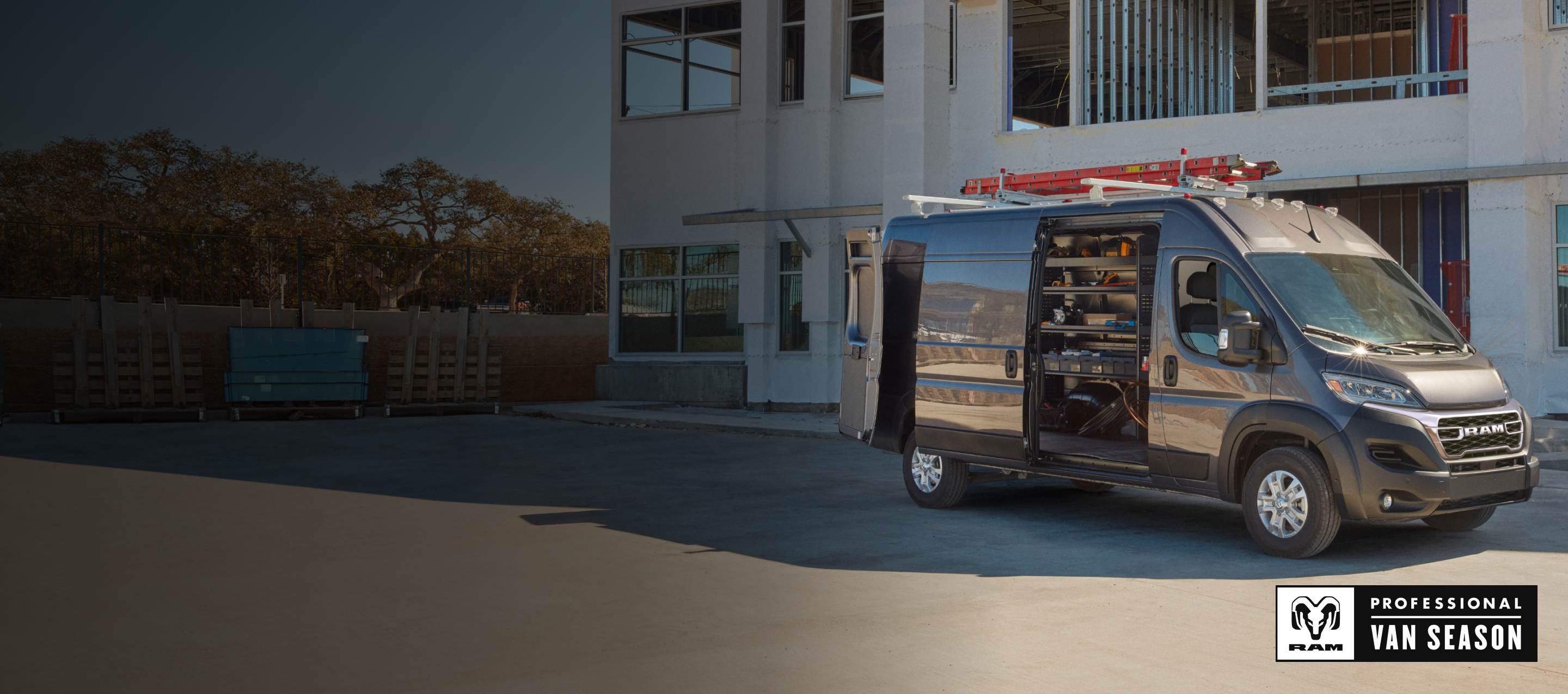 A dark gray 2025 Ram ProMaster 2500 SLT High Roof Cargo Van parked beside a commercial building with a ladder strapped to its roof rack, its rear doors and sliding side door open revealing shelving full of tools. Ram Professional Van Season.
