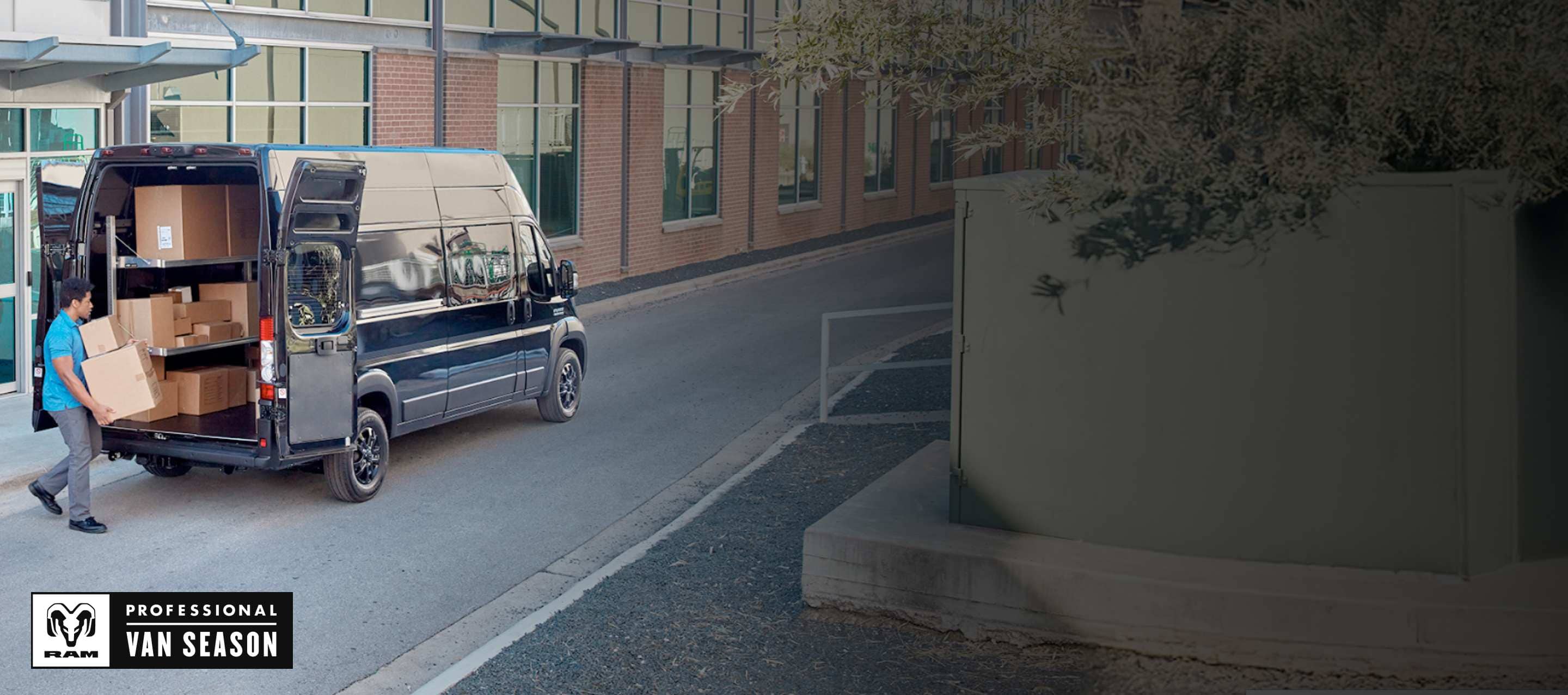 A man carrying packages toward the open rear doors of a black 2025 Ram ProMaster 3500 SLT Plus Super High Roof Cargo Van with its cargo area shelves full of boxes. Ram Professional Van Season.