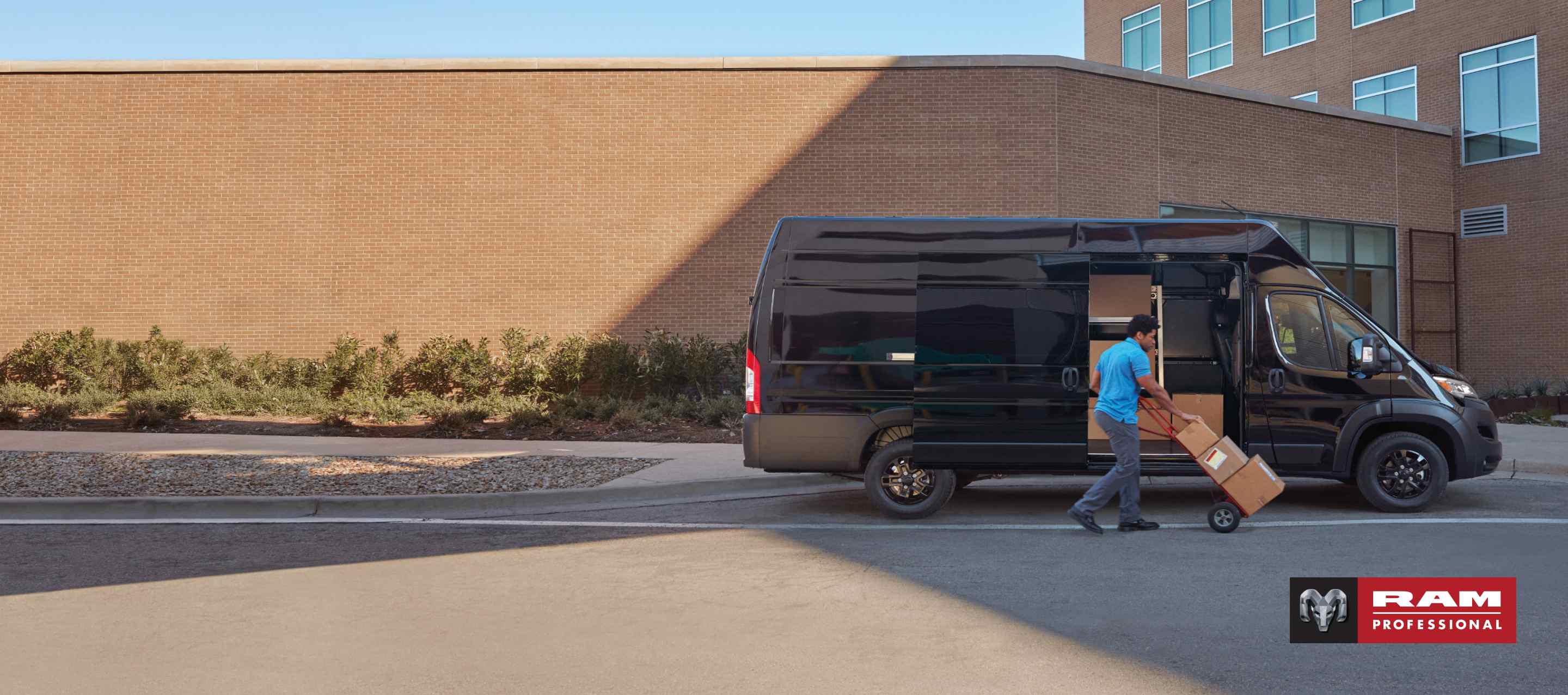 A man pushing packages on a dolly near the open passenger-side sliding door on a black 2025 Ram ProMaster 3500 SLT Plus Super High Roof Cargo Van with the Black Appearance Package. Ram Professional.