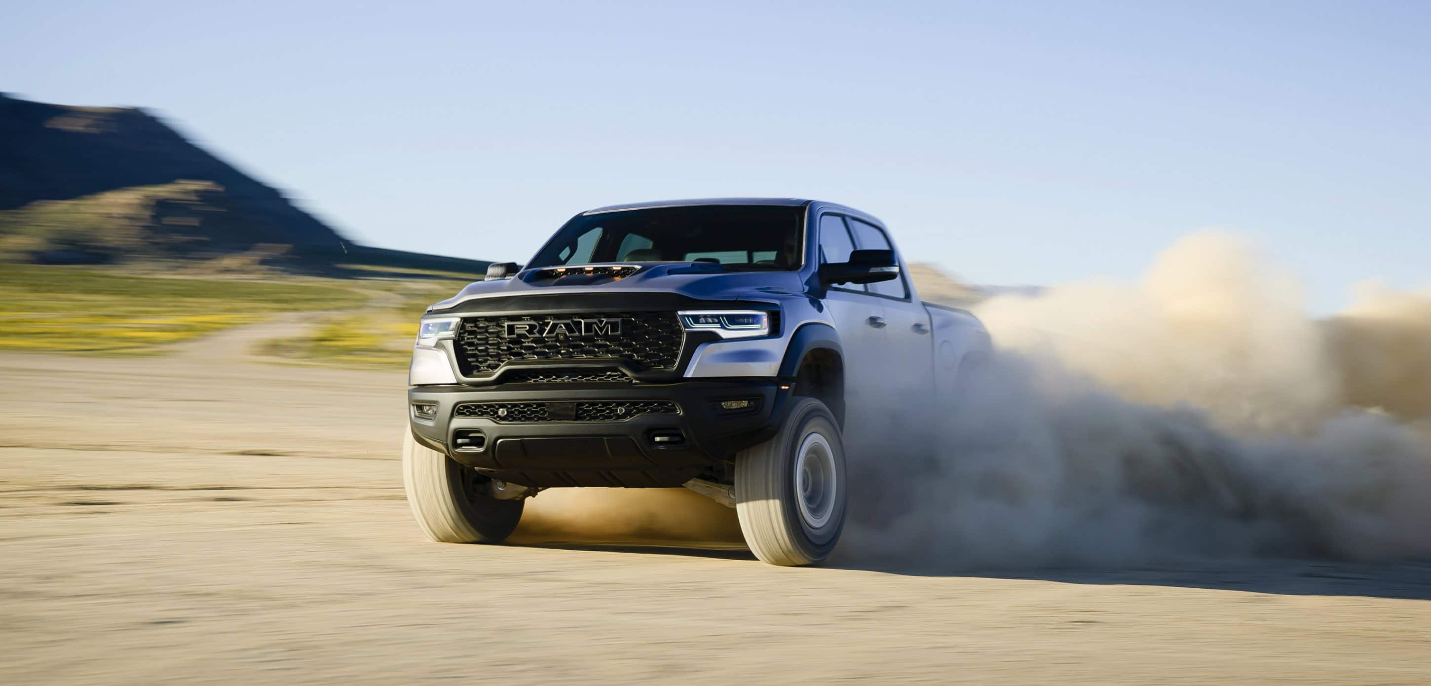 An angled driver-side front profile of a silver 2025 Ram 1500 RHO Crew Cab being driven off-road in the desert, with dust and sand billowing from its tires.