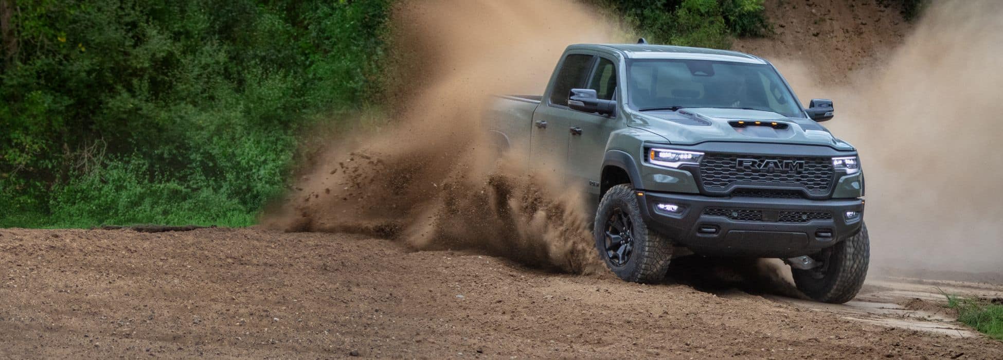 A bluish-silver 2026 Ram 1500 RHO 4x4 Crew Cab turning on a sandy trail off-road, kicking up a cloud of dust as it goes.