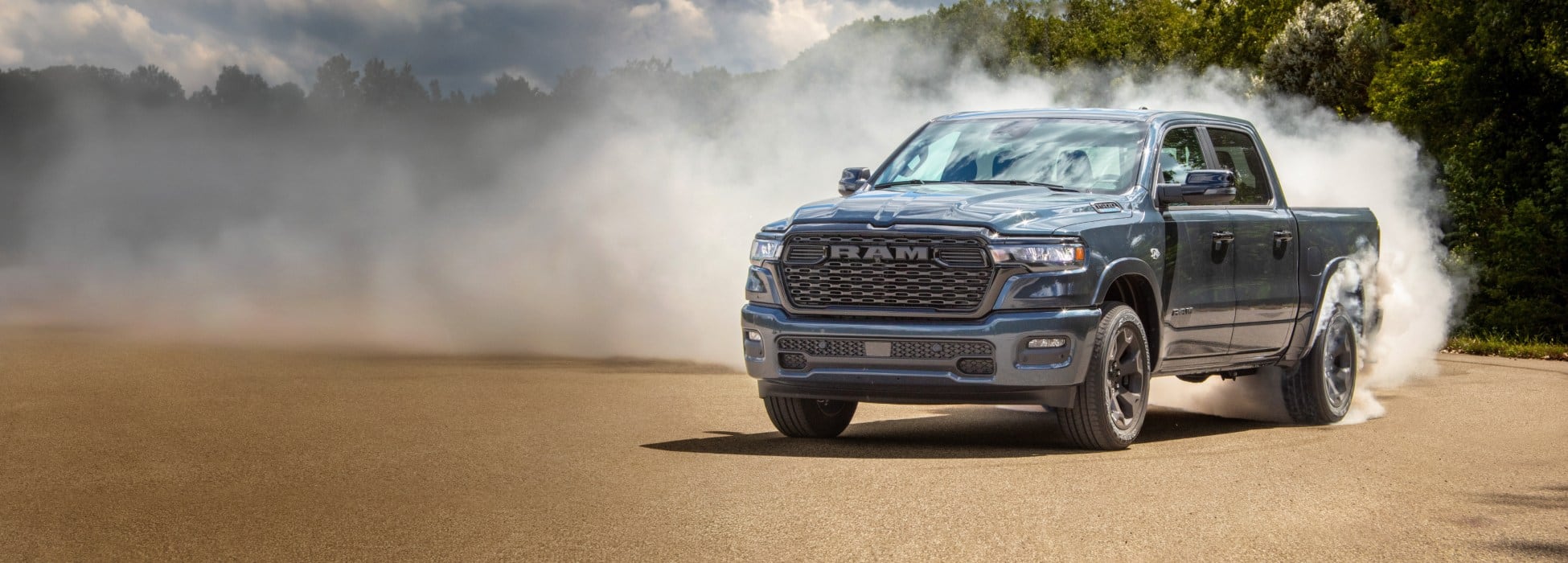 A bluish-silver 2026 Ram 1500 Big Horn 4x4 Crew Cab turning on a sandy trail off-road, with white smoke billowing from its rear tires.