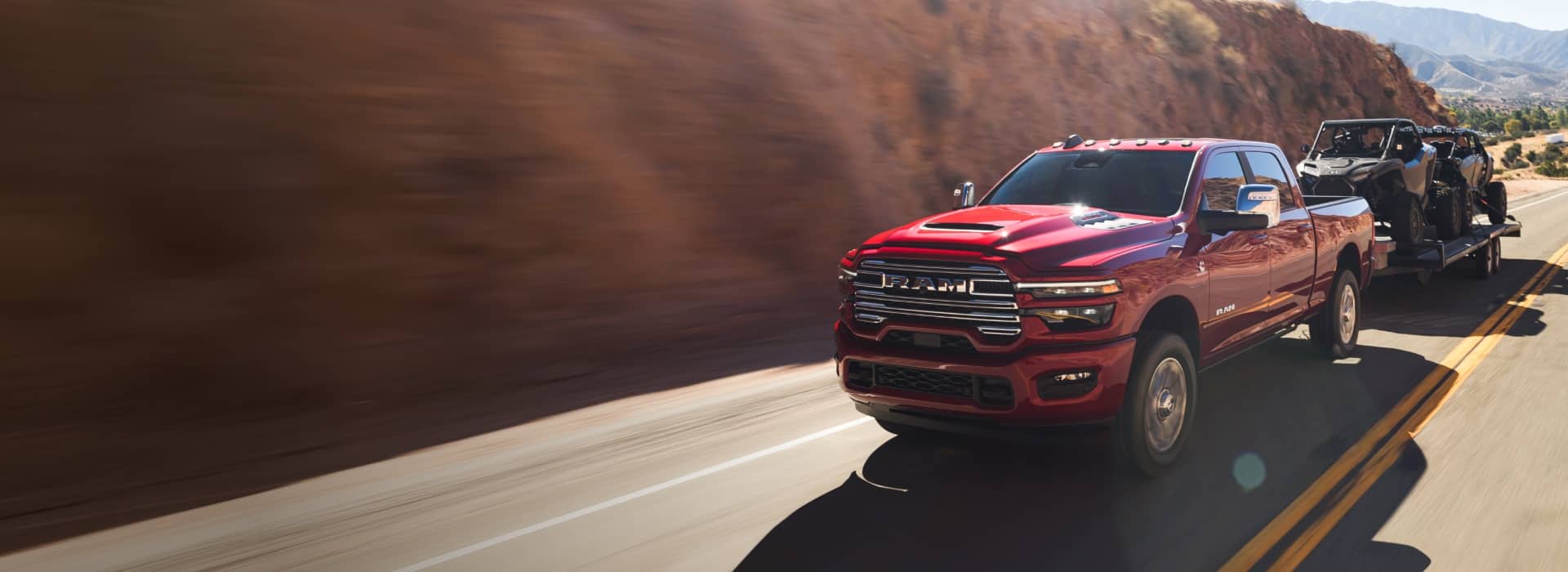 A red 2026 Ram 2500 Laramie 4x4 Crew Cab traveling on a highway in the mountains, while towing two ATVs.