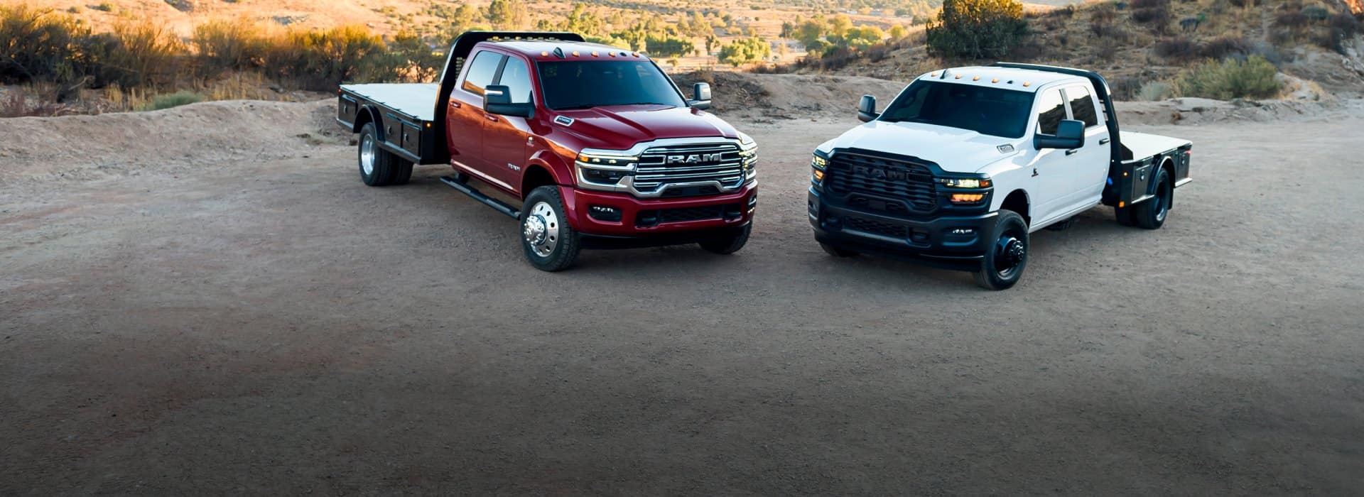 Two 2026 Ram Chassis Cabs, both with platform upfits, parked on a clearing. On the left, a red 2026 Ram 5500 Laramie Chassis Cab Crew Cab and on the right, a white 2026 Ram 3500 Tradesman Chassis Cab Crew Cab.