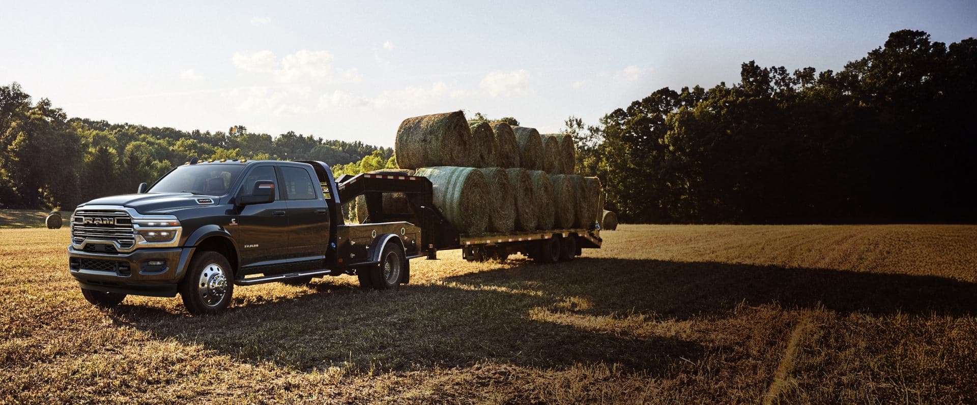 A black 2026 Ram 5500 Laramie Chassis Cab Crew Cab with a gooseneck trailer loaded with several bales of hay, parked in a field.