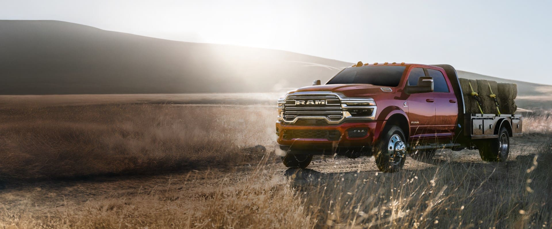 A red 2026 Ram 5500 Laramie Chassis Cab Crew Cab carrying several large bales of hay on a long platform upfit as it travels through ranch land.