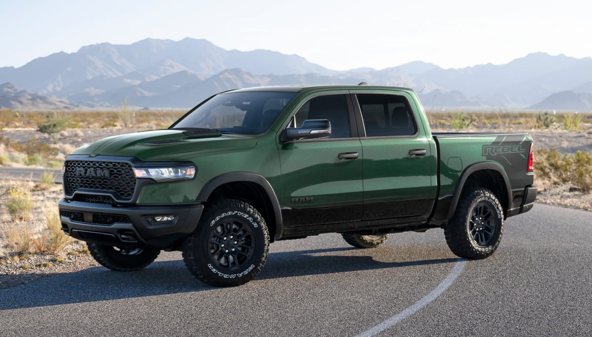 Display A green 2026 Ram 1500 Rebel 4x4 Crew Cab parked on a desert road with mountains in the background.