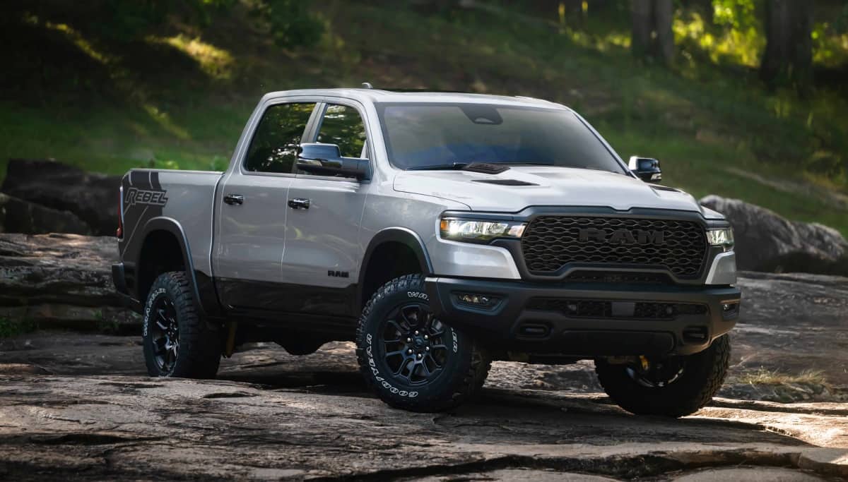 Display A white 2026 Ram 1500 Rebel 4x4 Crew Cab traveling down a rocky trail off-road, with a forest in the background.